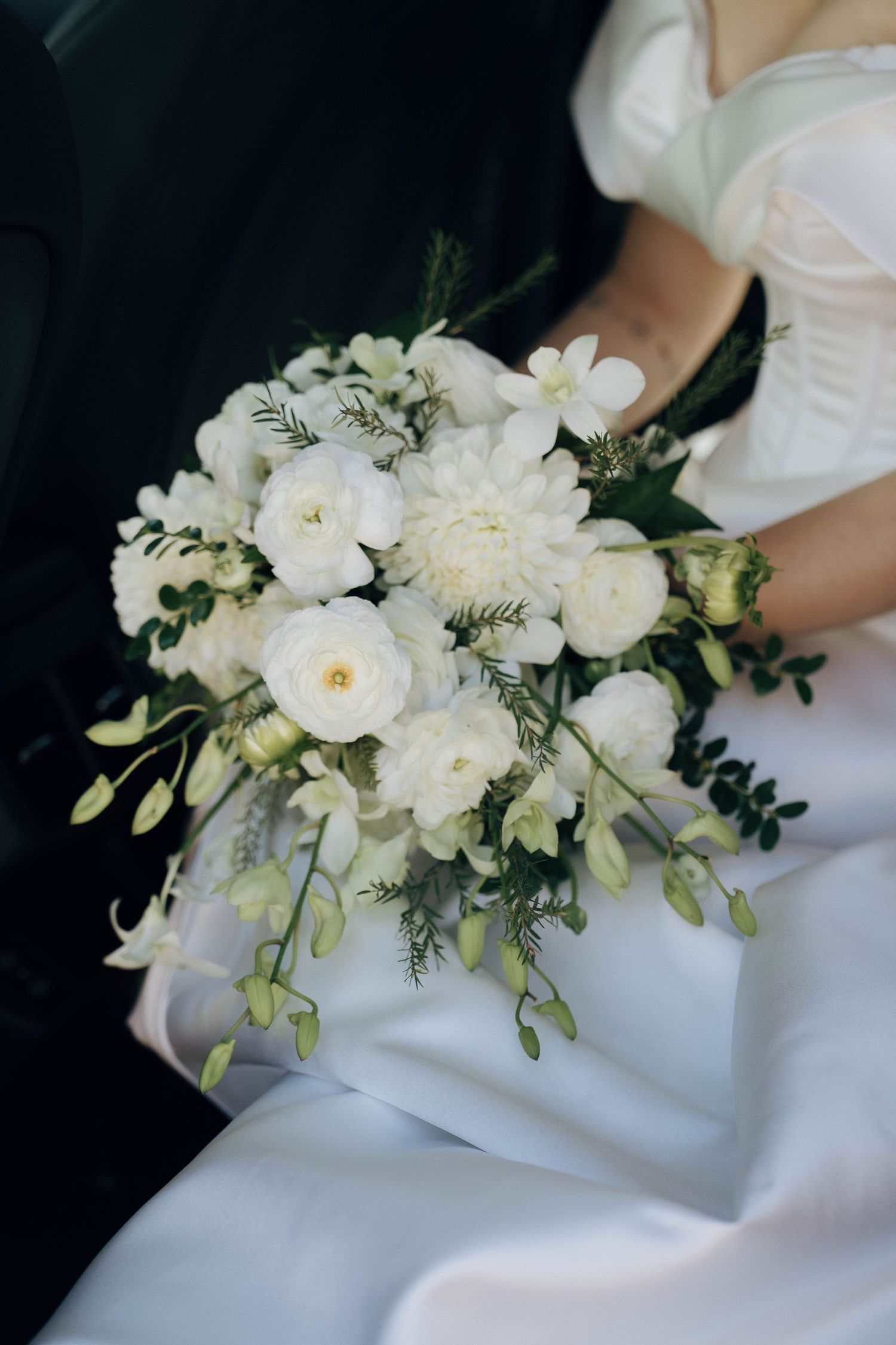 A delicate white floral wedding bouquet with greenery rests elegantly against a white dress.
