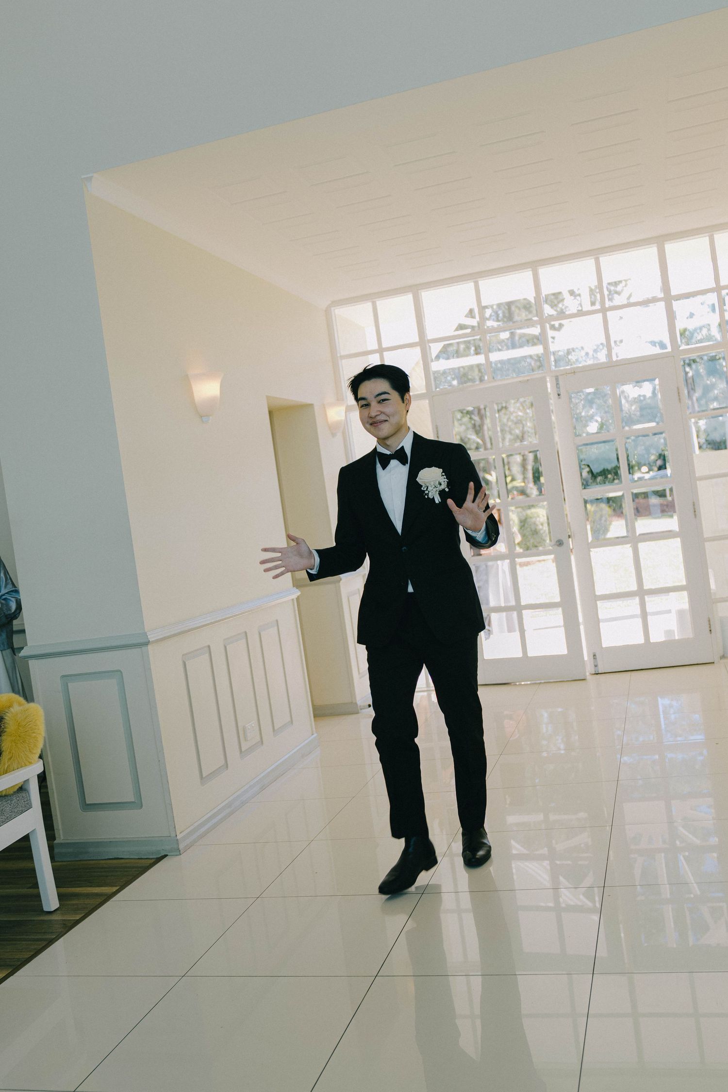 A person in a black suit gestures expressively while walking through a bright, modern hallway with white floors and walls.