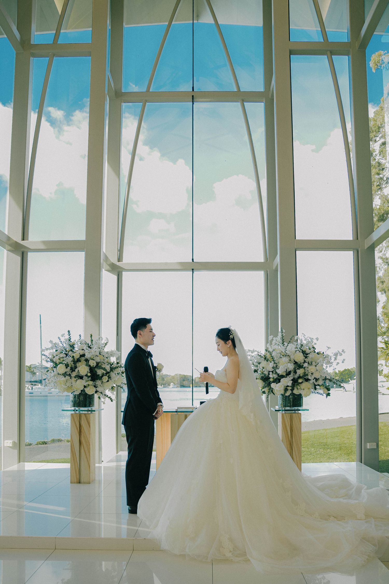 A panoramic view of a modern glass chapel wedding ceremony with white floral arrangements and water views in the background.