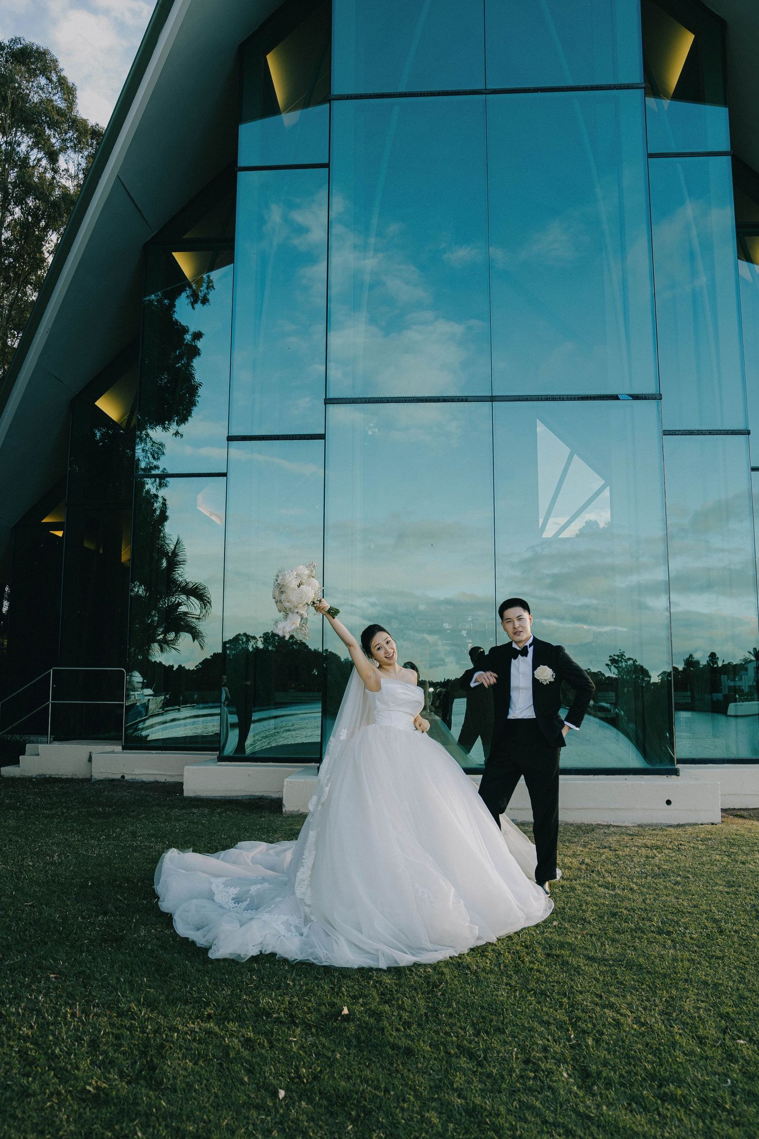 A couple in wedding attire poses together on a grassy hill against a modern glass building exterior at sunset.