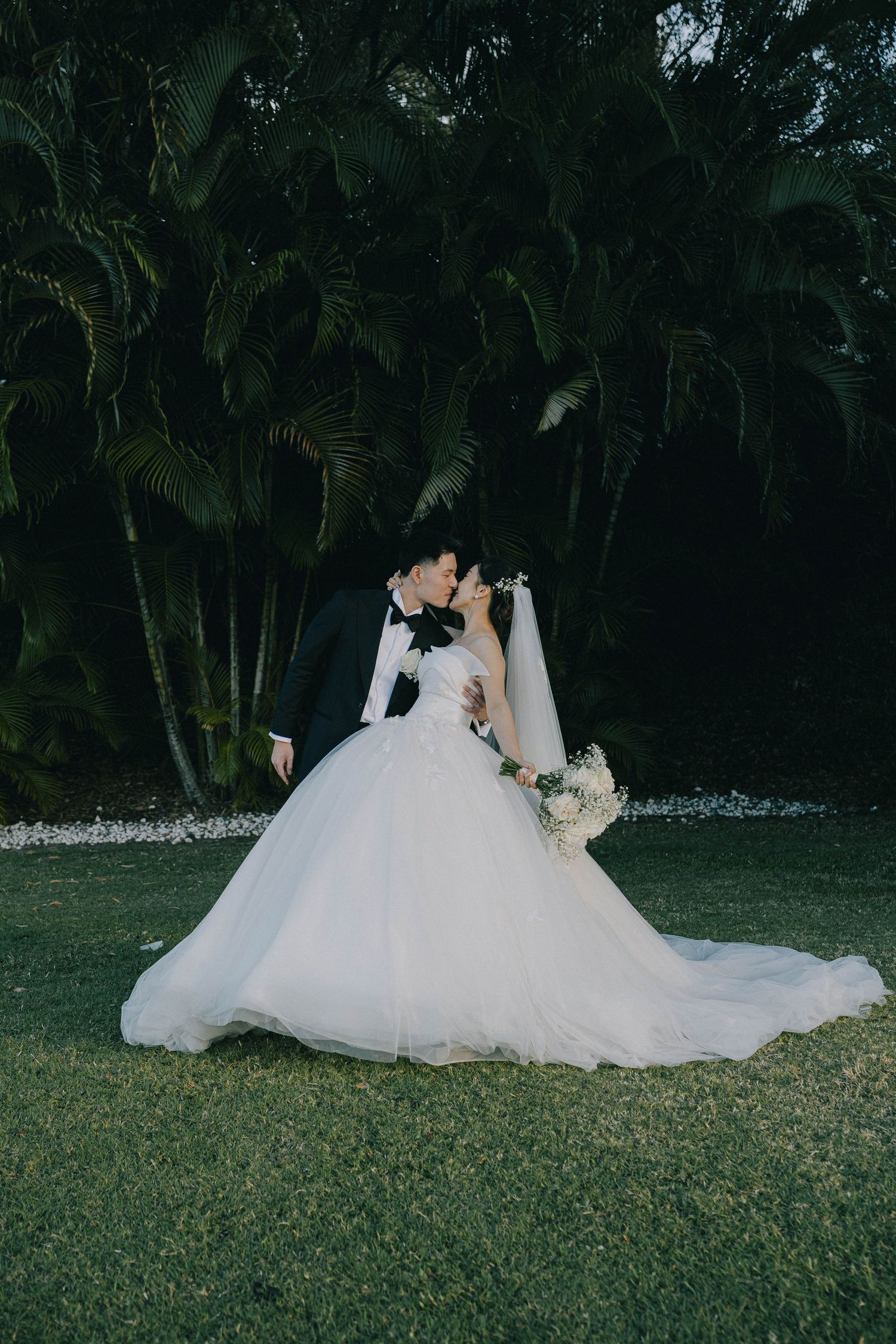 A bride in a flowing white ballgown wedding dress poses gracefully on a lush green lawn at dusk.