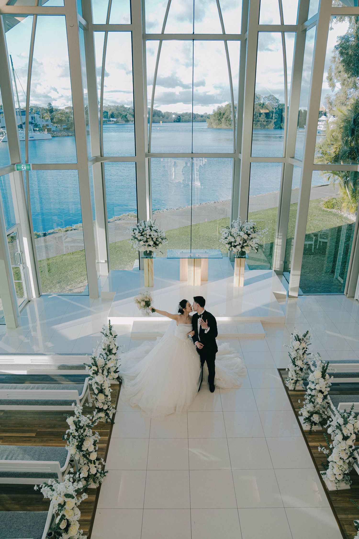 Modern glass chapel interior with white marble floors and floral arrangements creates an elegant wedding ceremony setting.