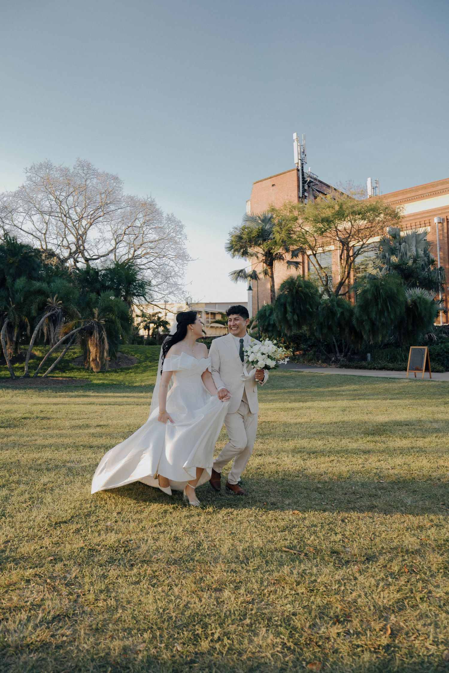 A couple in flowing white attire dances gracefully across a sunlit grassy field with buildings in the background.