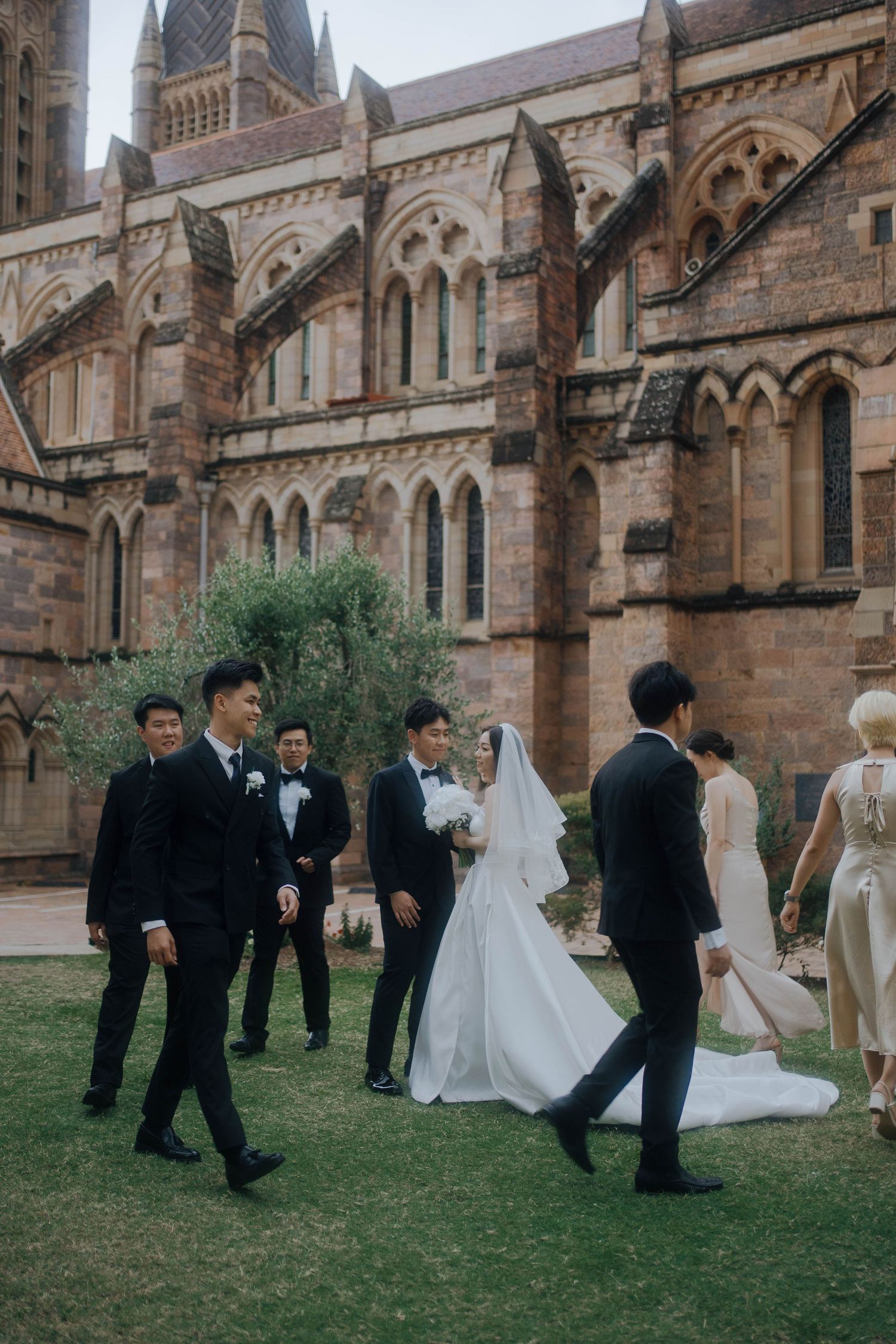Wedding party poses on grass lawn outside historic gothic church building in elegant formal attire.