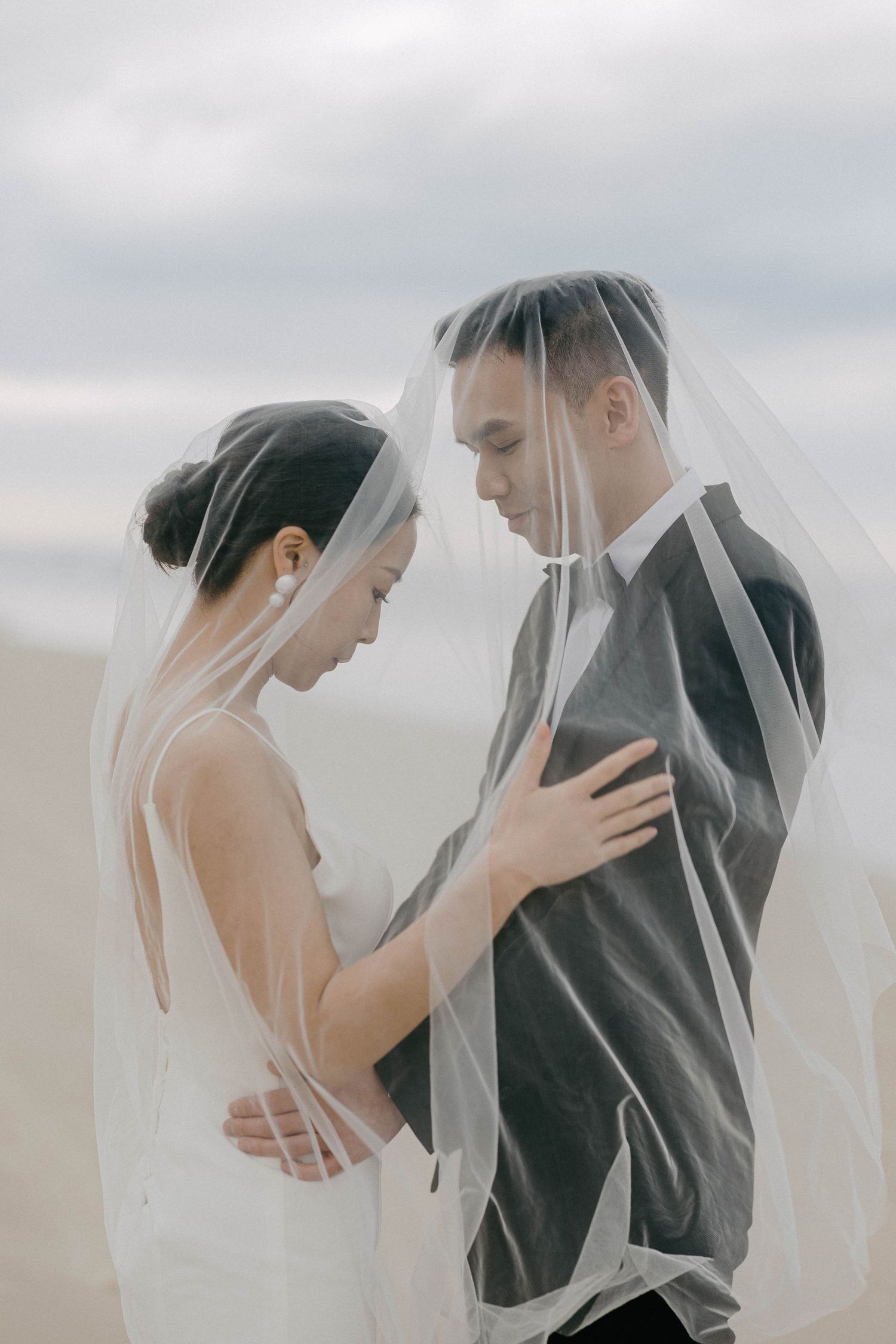 A romantic wedding couple embraces under a flowing veil against an overcast sky.