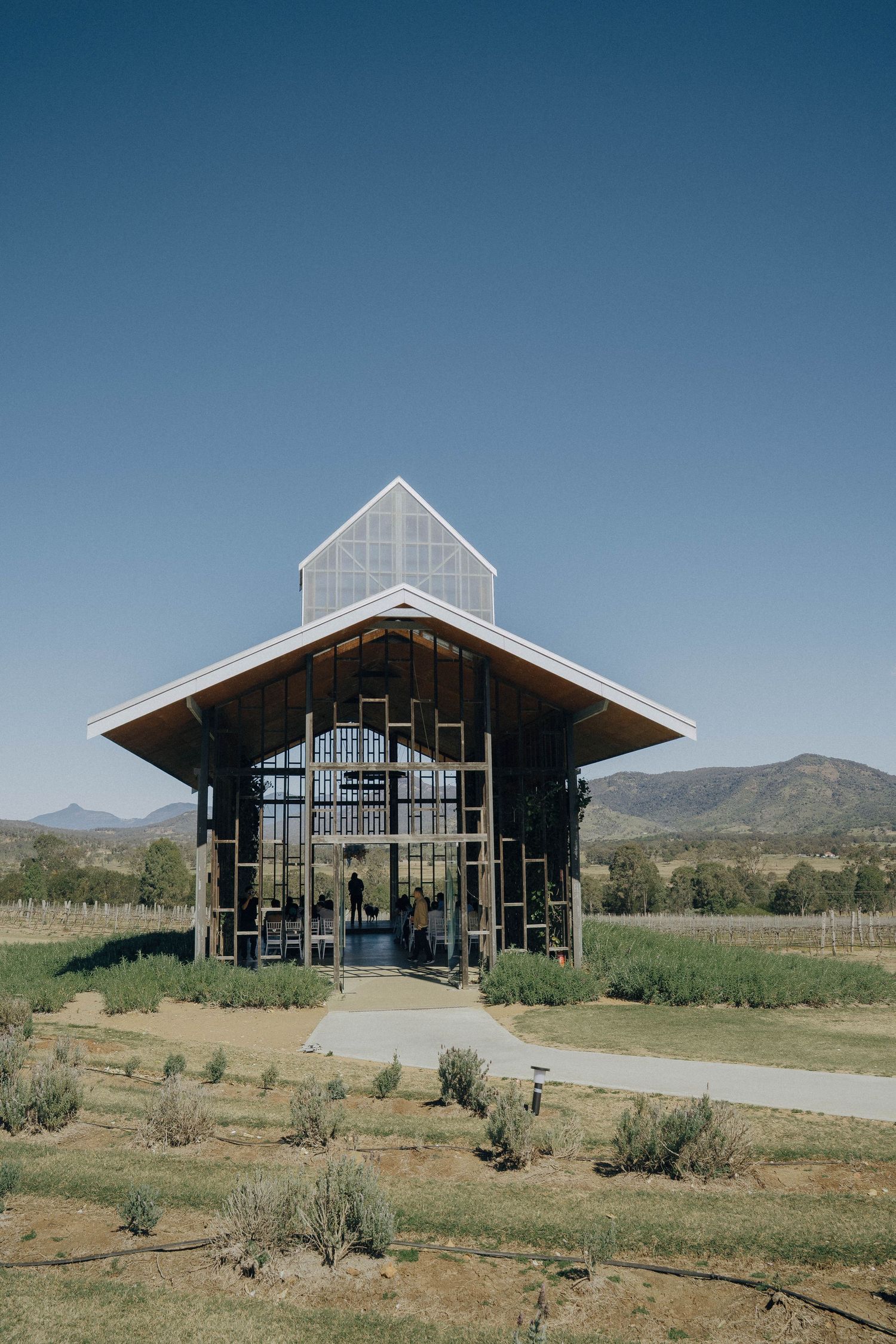 Modern rustic barn-style building with glass walls and angular roof sits in a scenic mountain landscape with desert brush.