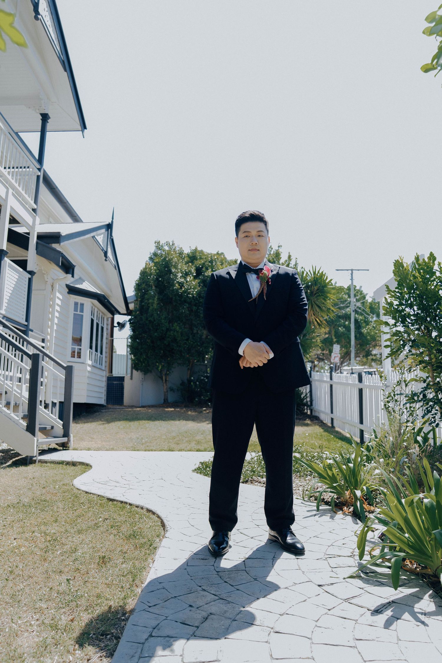 A person in a black tuxedo and bow tie stands on a curved stone pathway in front of a white house.
