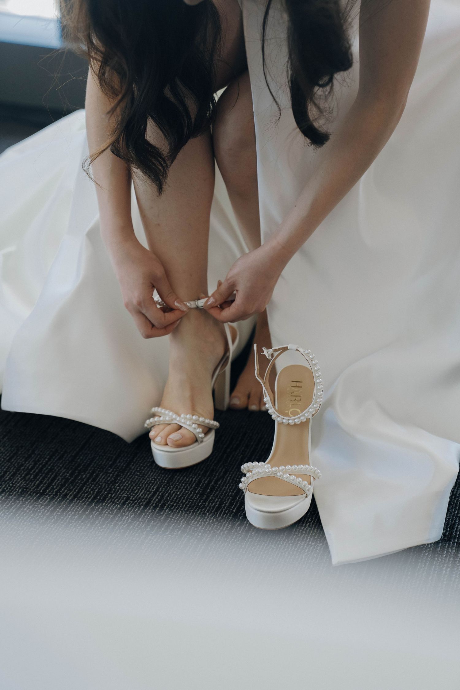 Close-up sequence of hands fastening delicate pearl-adorned white bridal shoes on carpeted floor.