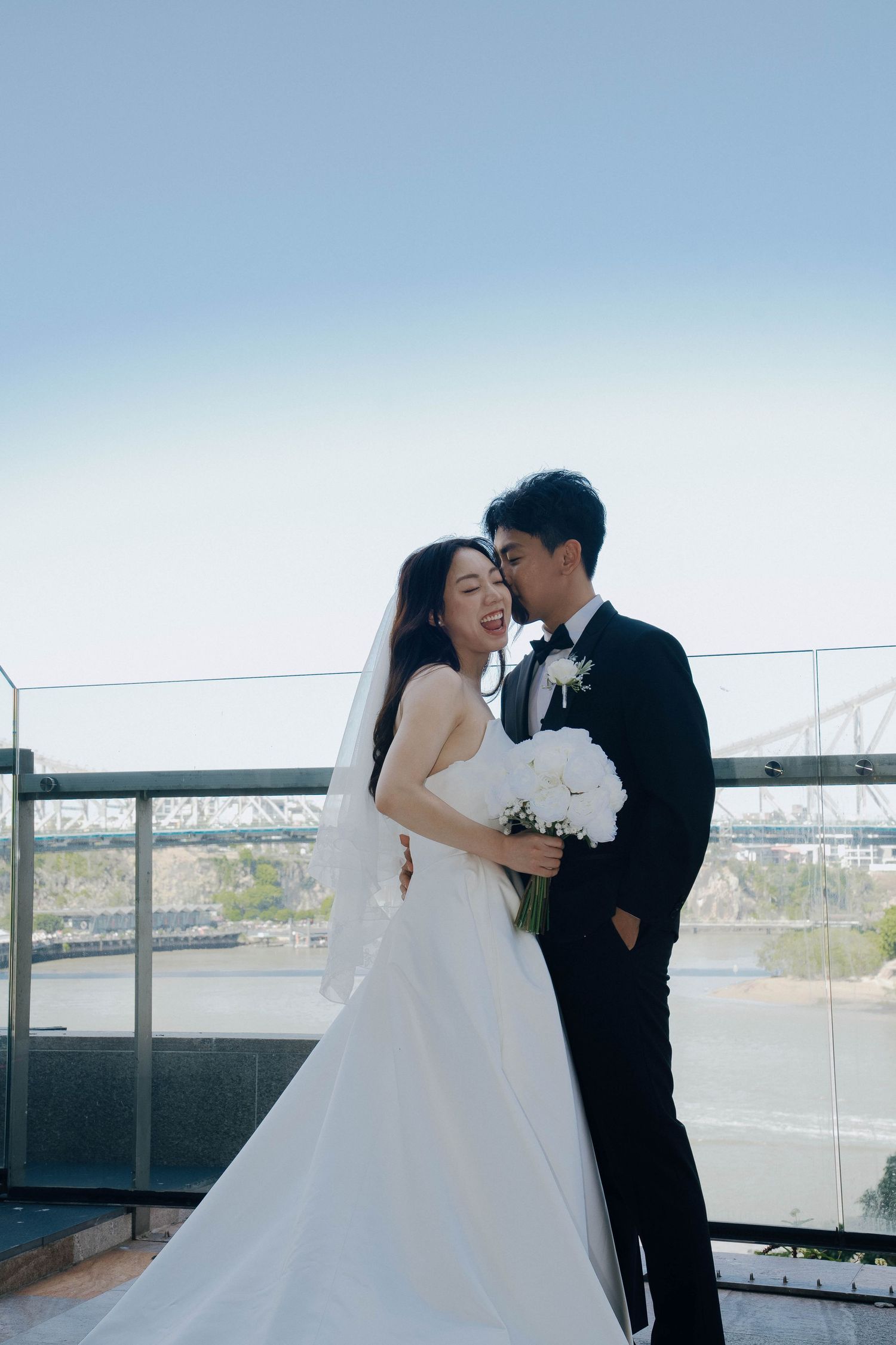 A romantic wedding couple shares a tender moment on a rooftop terrace overlooking a scenic waterfront view.