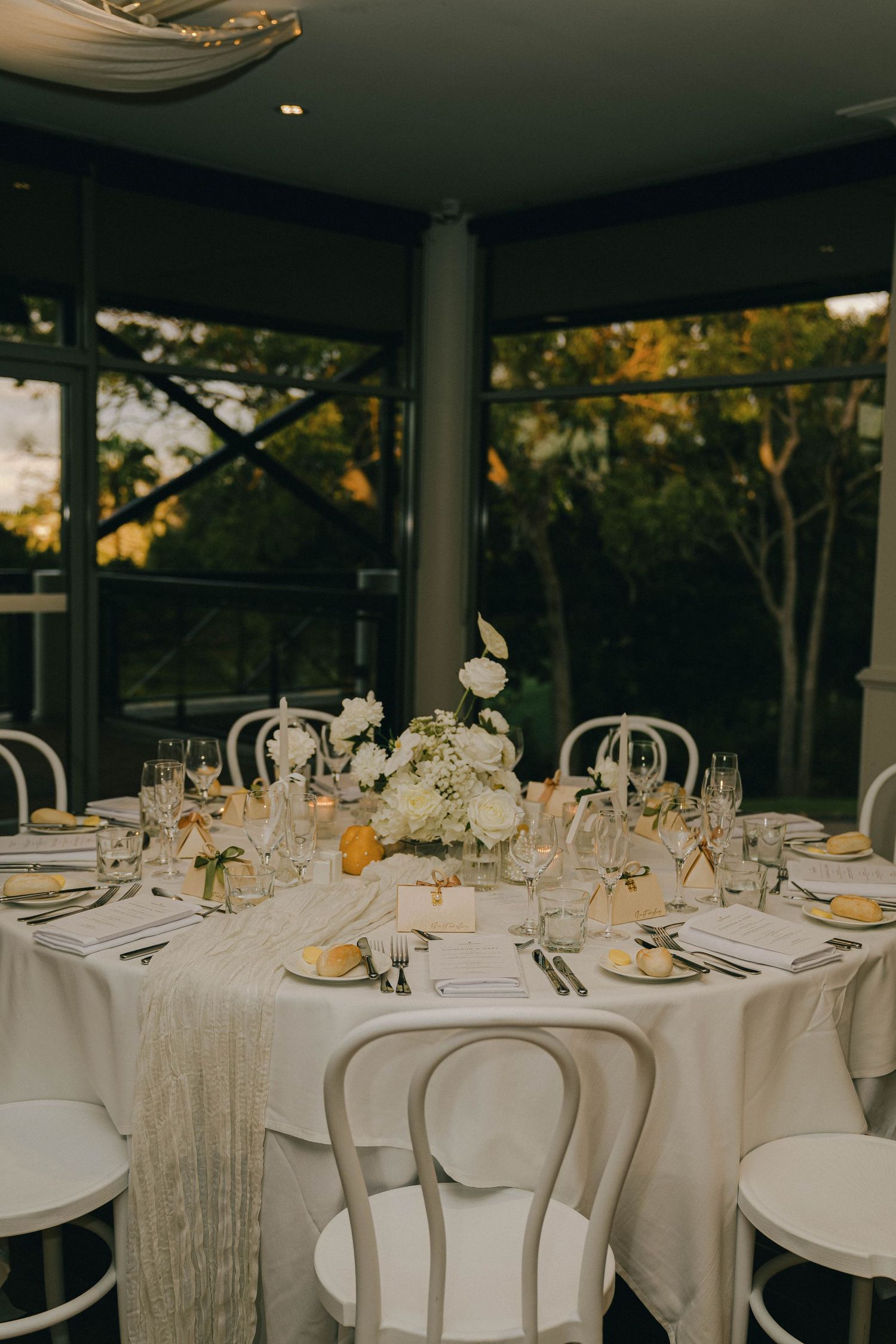 Elegant wedding reception tables set with white linens, glassware and floral centerpieces in a bright windowed venue.