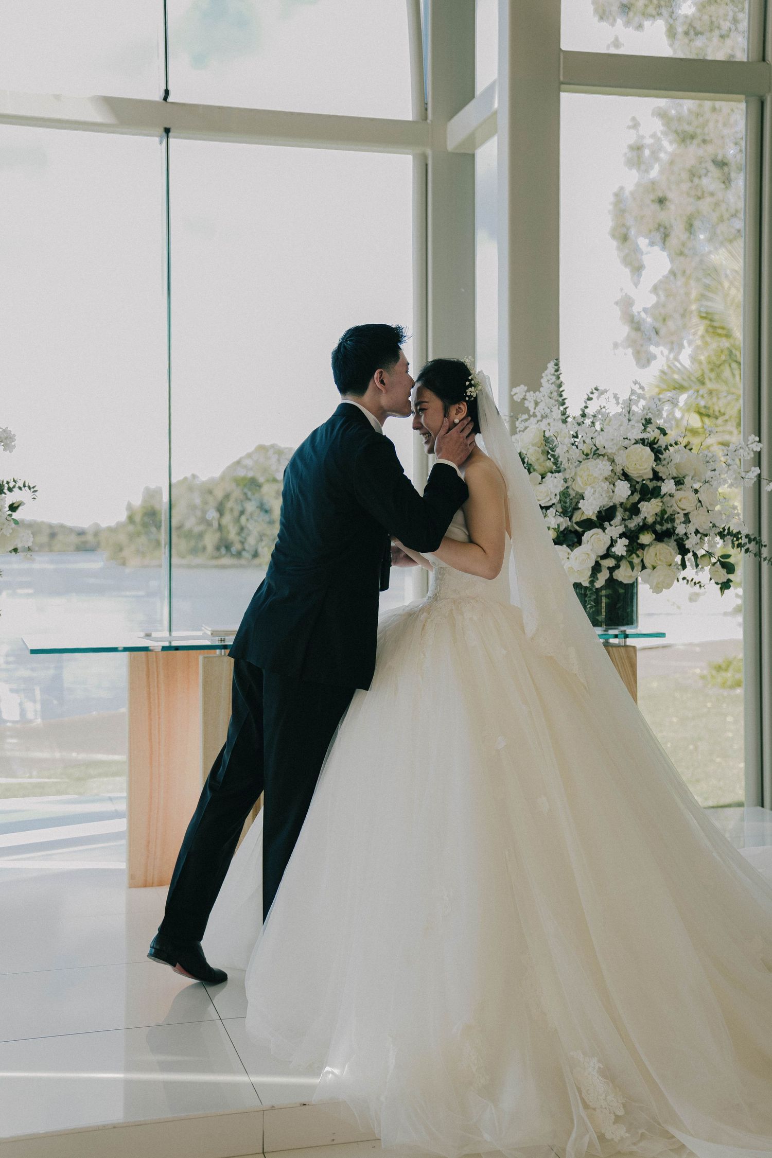 A couple in wedding attire share a romantic kiss in a bright, modern venue with large windows and white floral arrangements.