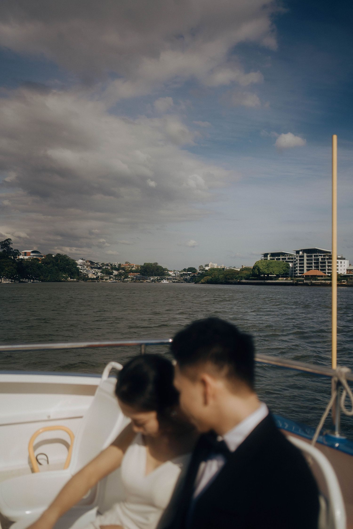 A couple shares an intimate moment on a boat with a scenic waterfront cityscape in the background.