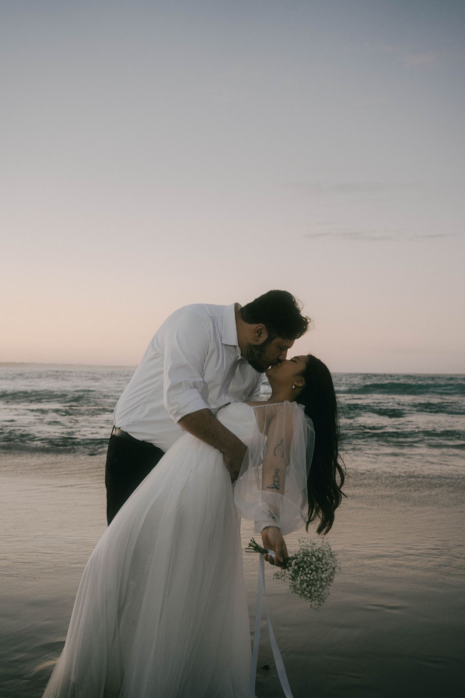 A romantic beach wedding photoshoot with a couple in white attire silhouetted against the ocean at sunset.
