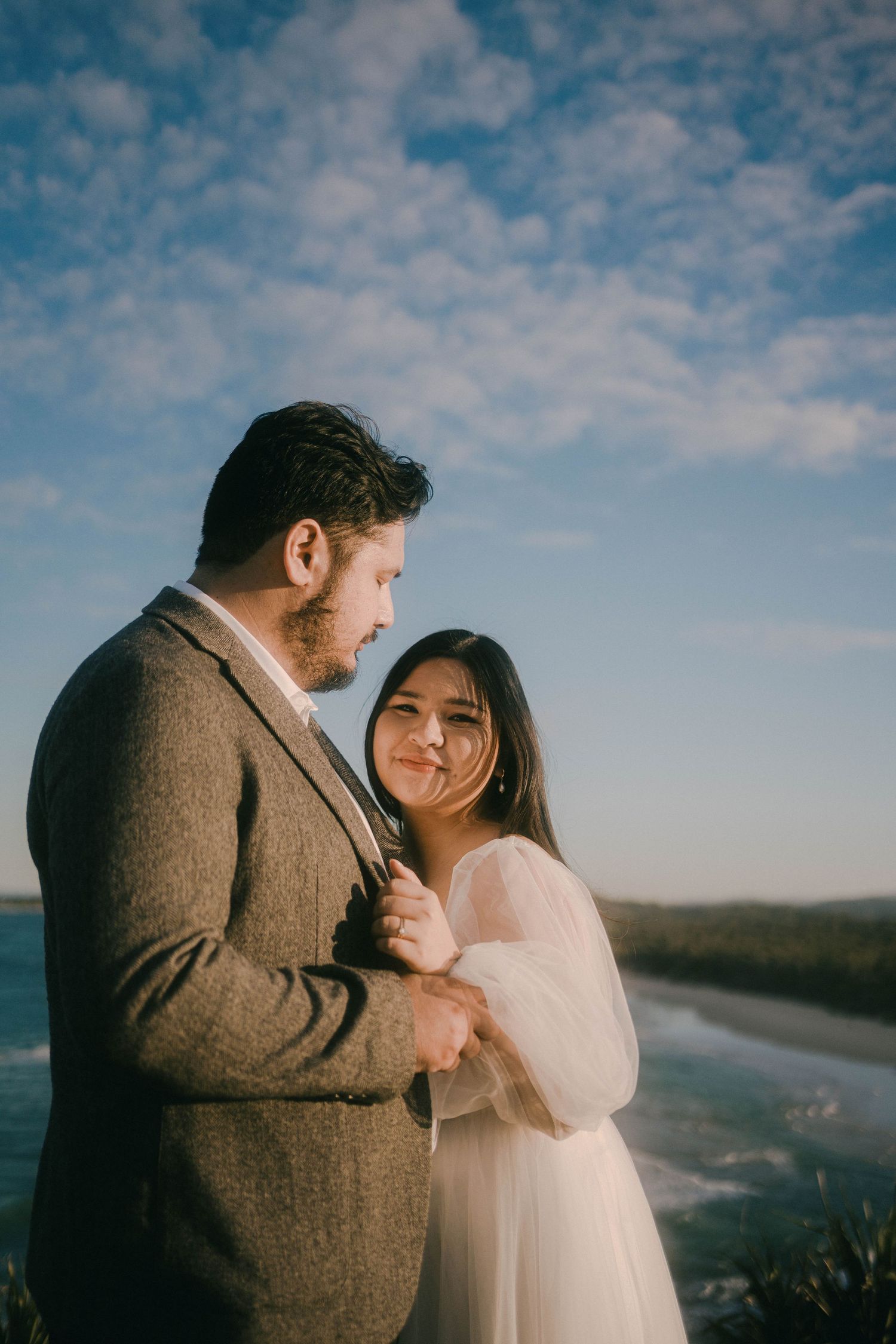 A romantic couple shares tender moments against a scenic coastal backdrop with dramatic cloudy skies.
