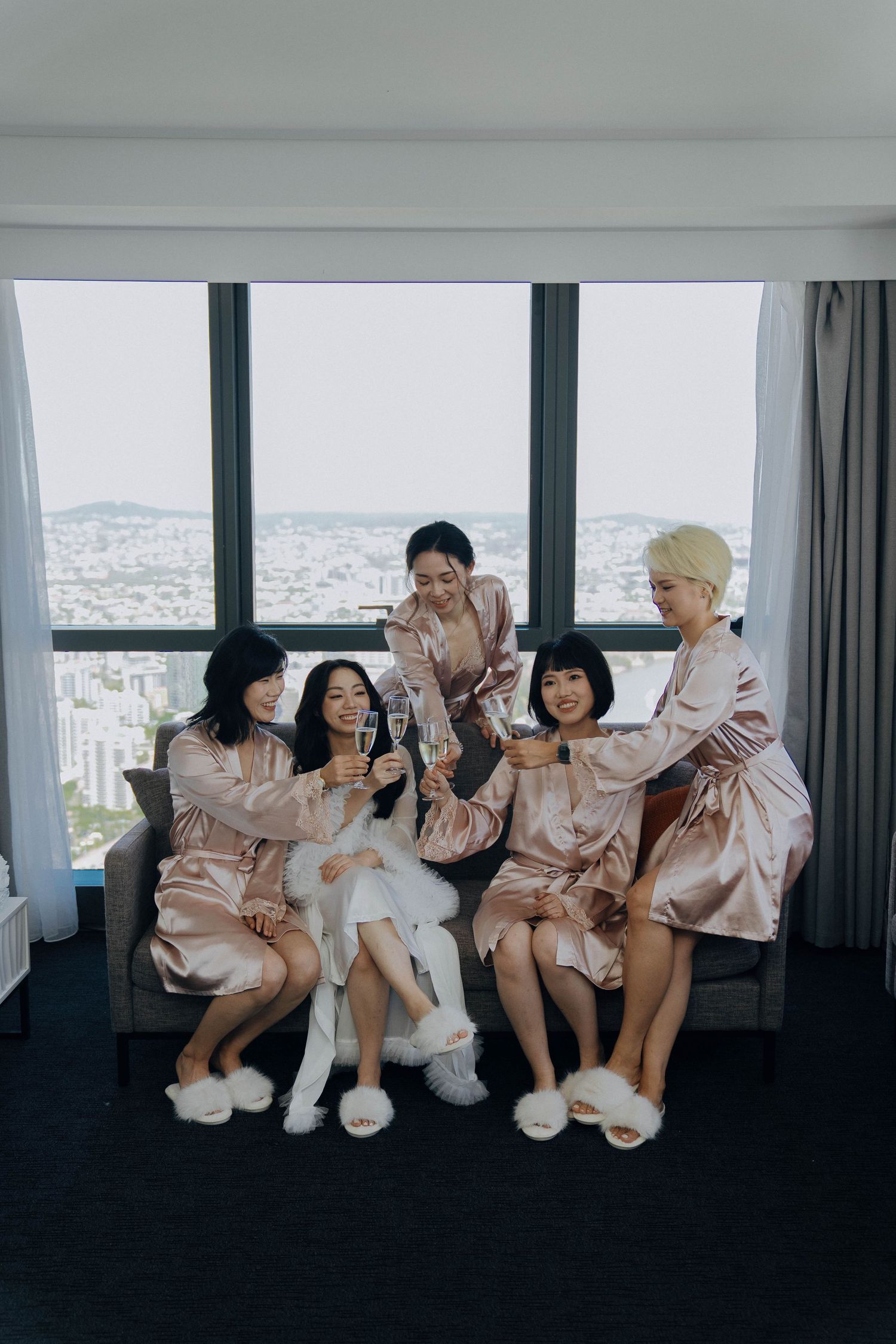 A group in matching outfits poses playfully on a couch against floor-to-ceiling windows overlooking a cityscape.