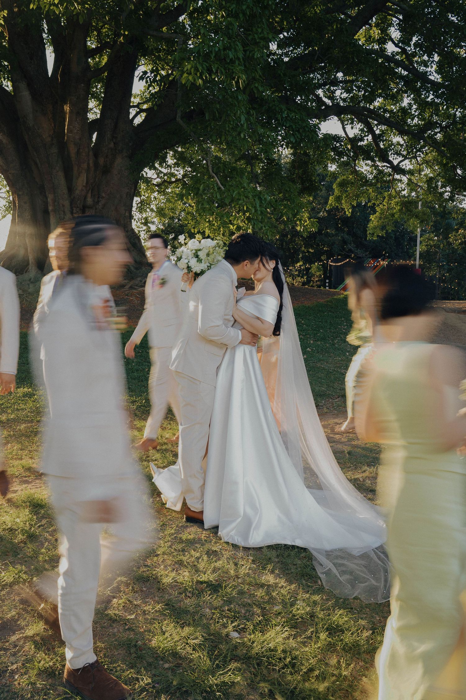 Wedding party in white attire walks through a sunlit garden setting creating dreamy motion blur effect.