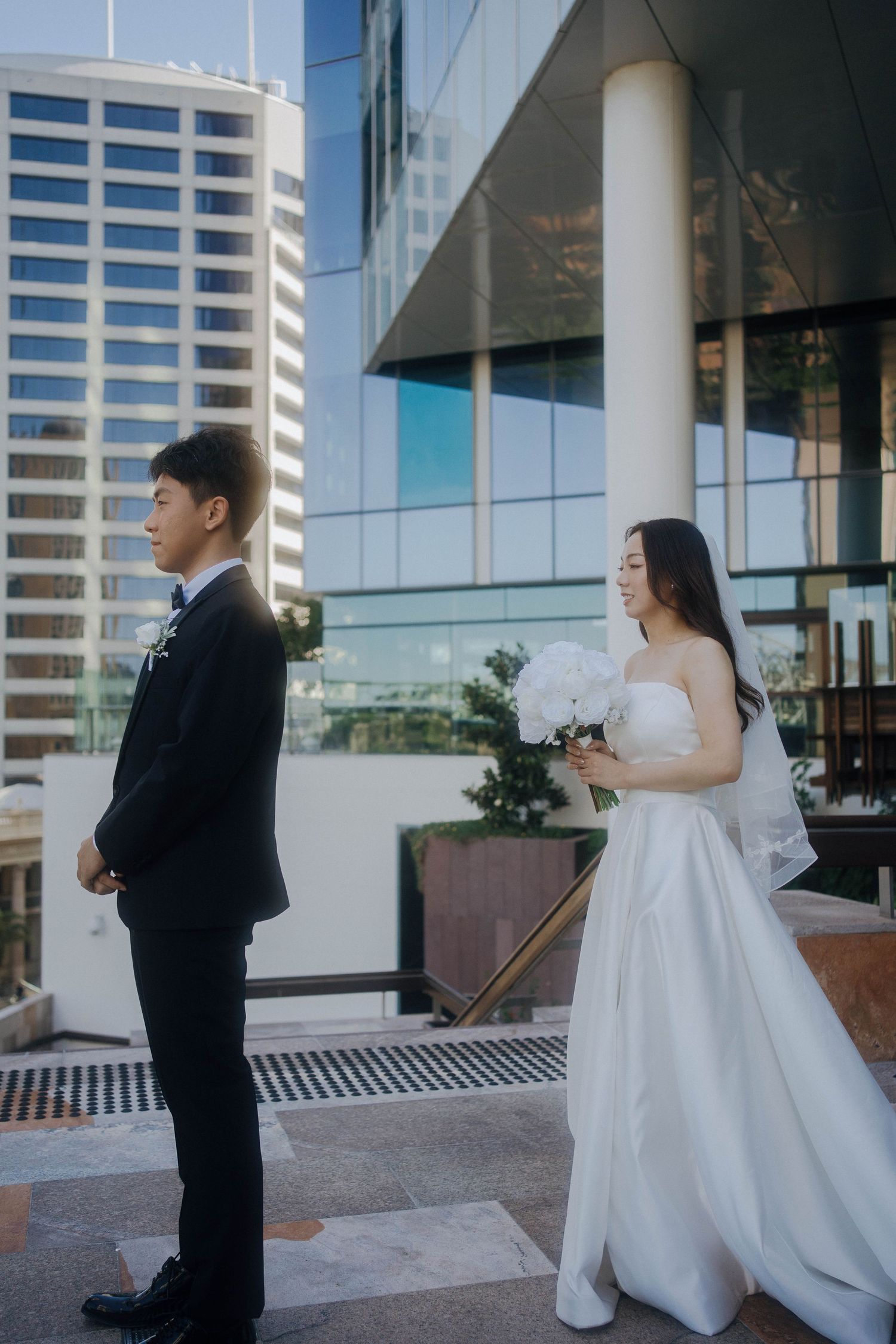 A couple in formal wedding attire shares tender moments outside a modern glass building.