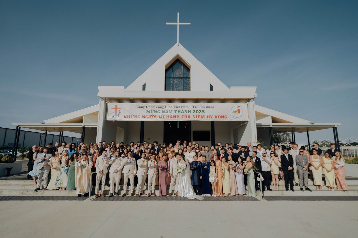 A modern white church building with a cross on top stands against a blue sky as wedding guests line up outside.