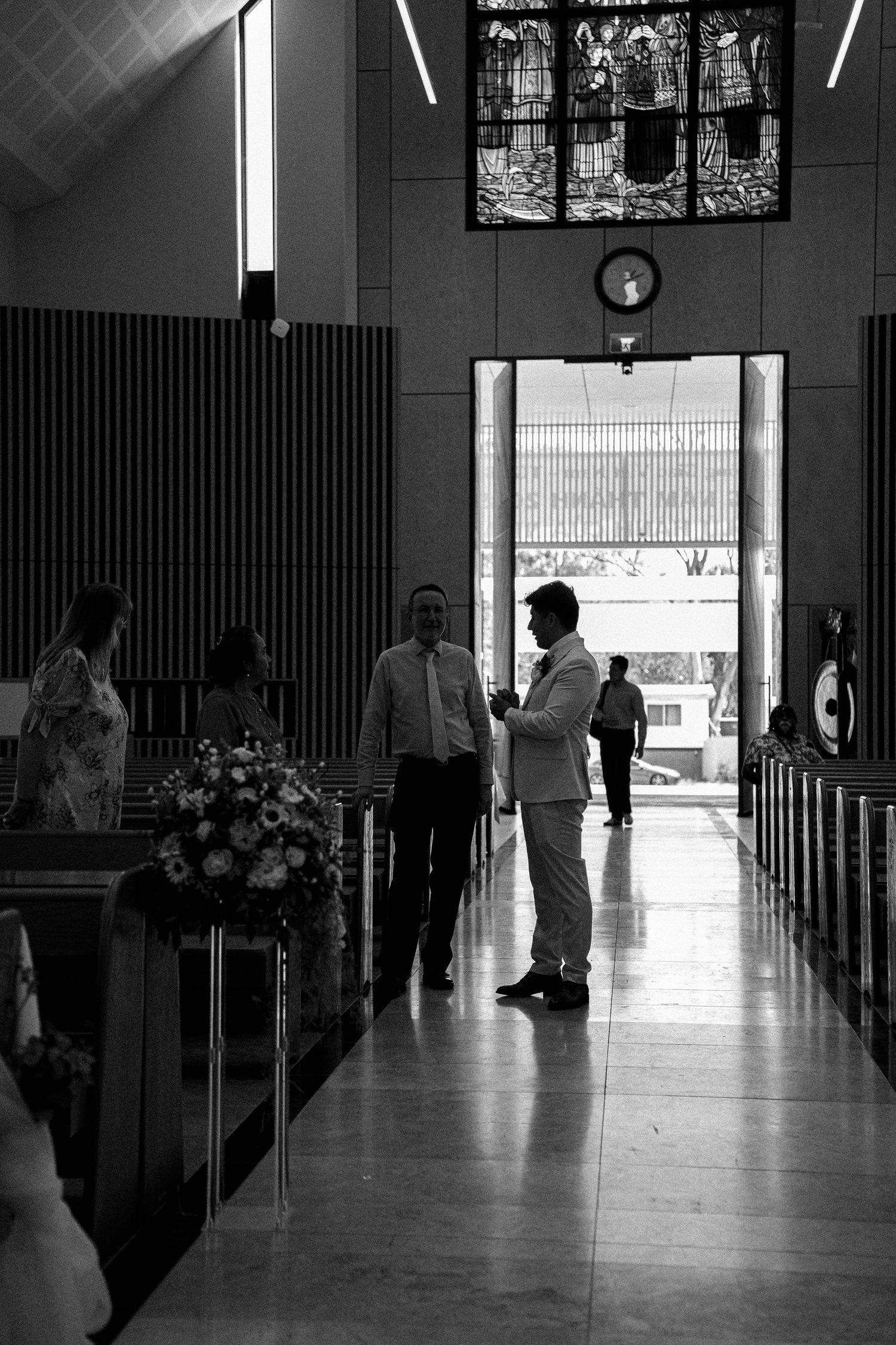 A black and white image shows silhouettes of people walking through a church hallway with bright light streaming in.