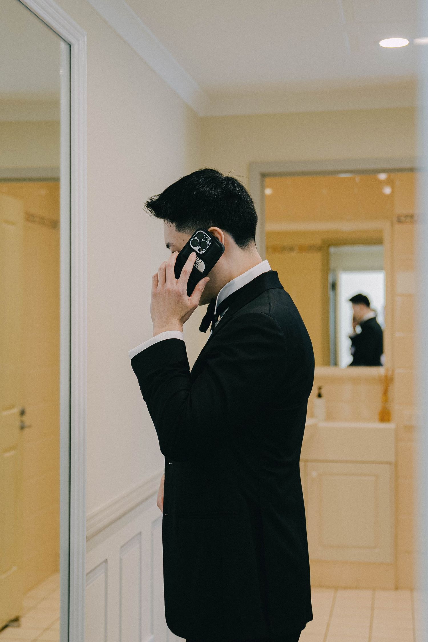 A person in a black suit speaks on a mobile phone in a hallway with cream colored walls and mirrors.