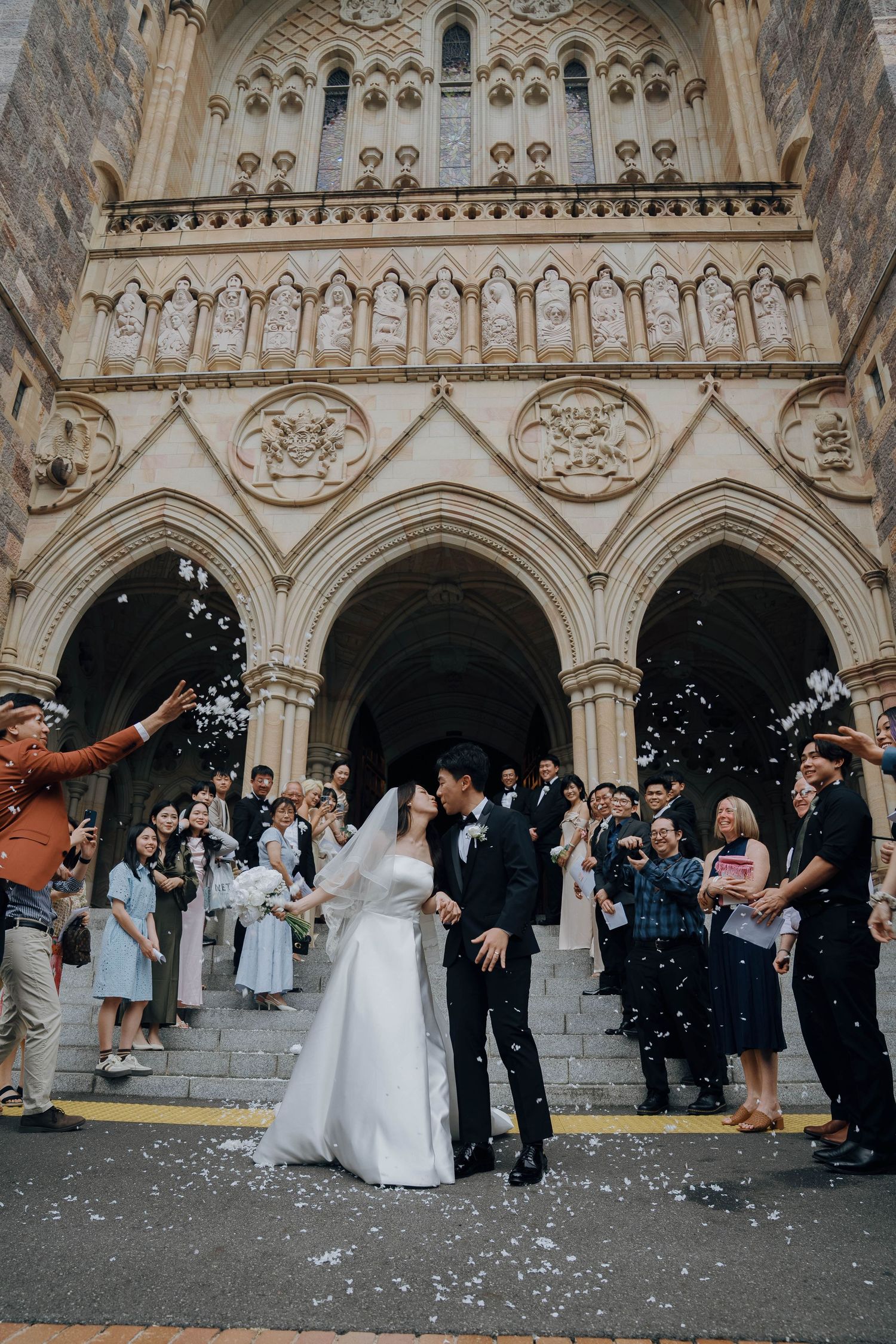 Wedding party exits church under shower of white confetti while guests celebrate on ornate Gothic cathedral steps.