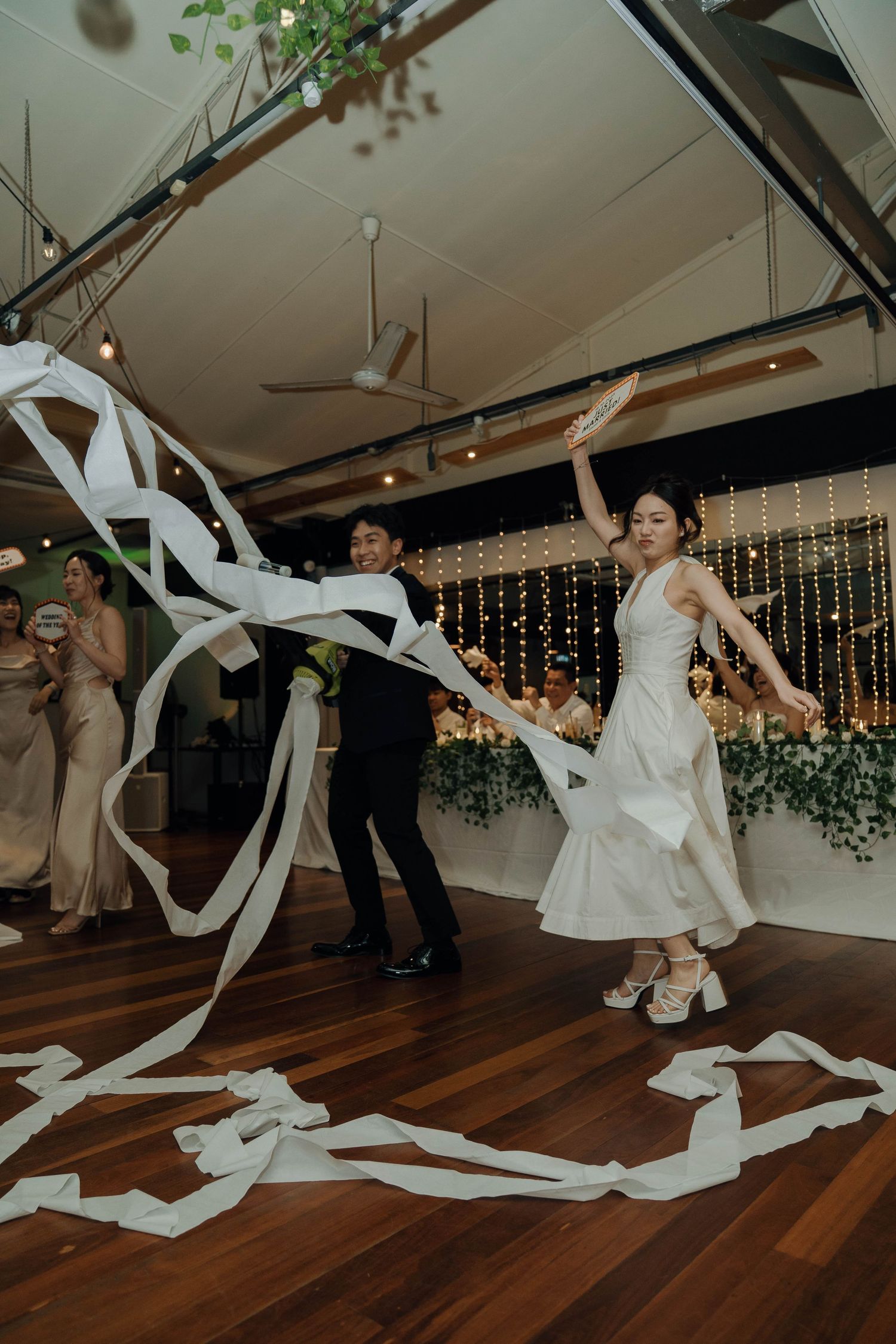Wedding guests dancing and twirling with long white streamers on a polished wooden dance floor at an indoor reception.