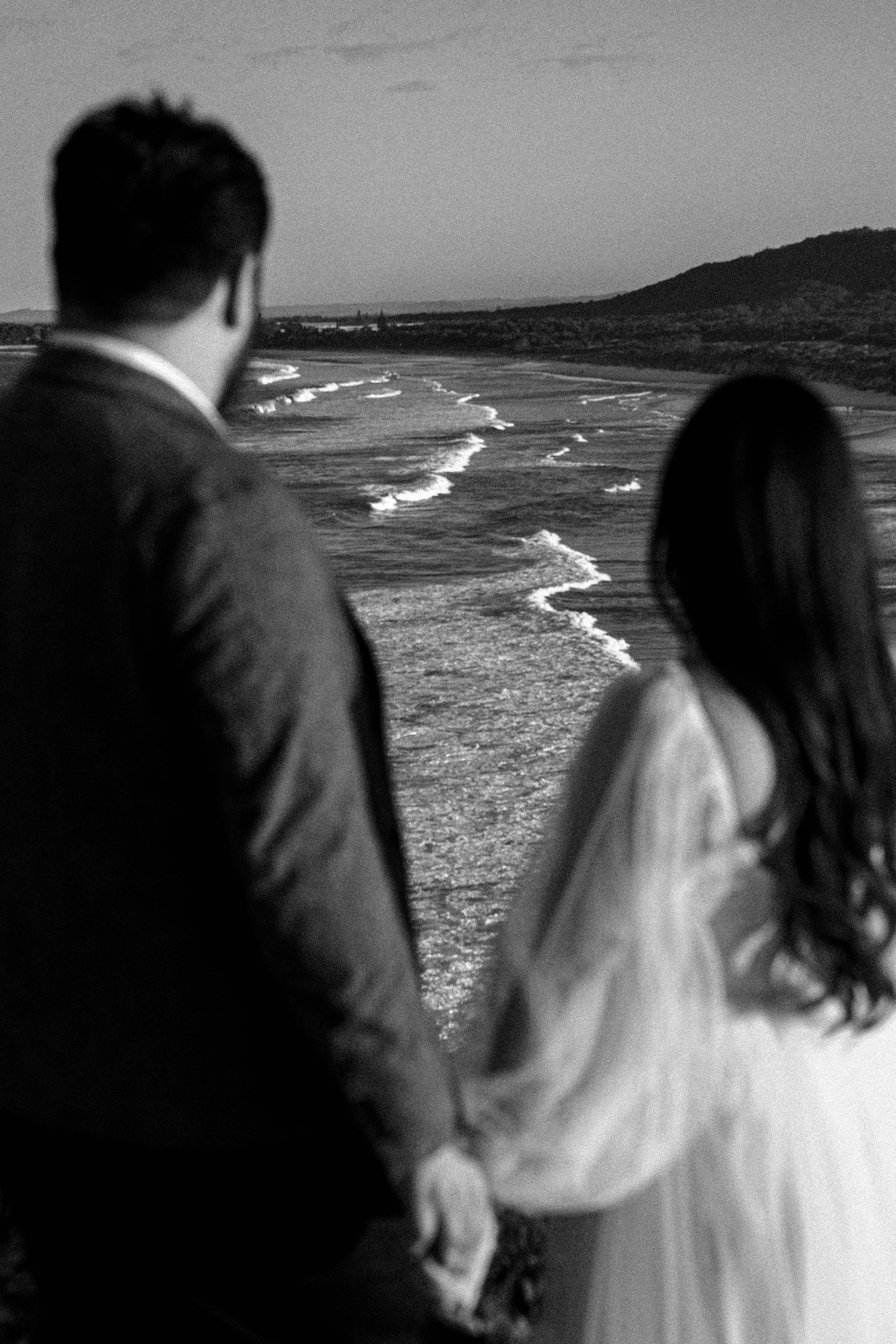 A couple holds hands while overlooking a scenic coastal landscape with crashing waves in black and white.