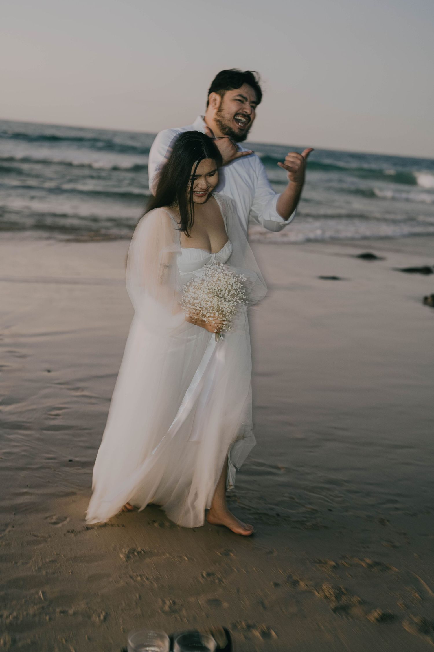 A couple in wedding attire shares an intimate moment on a moody beach with crashing waves in the background.