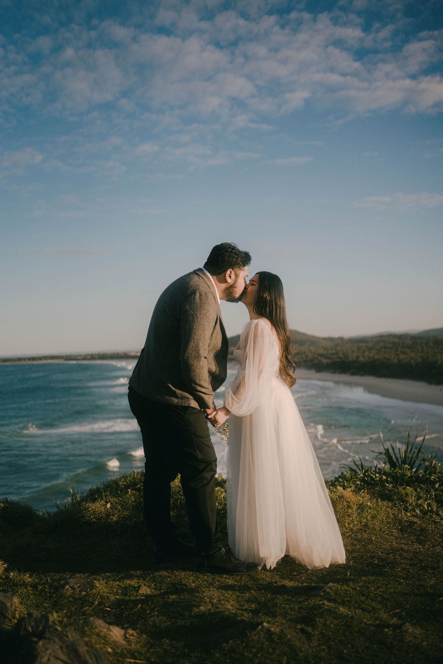 A couple in wedding attire shares an intimate moment overlooking a scenic coastal landscape at sunset.