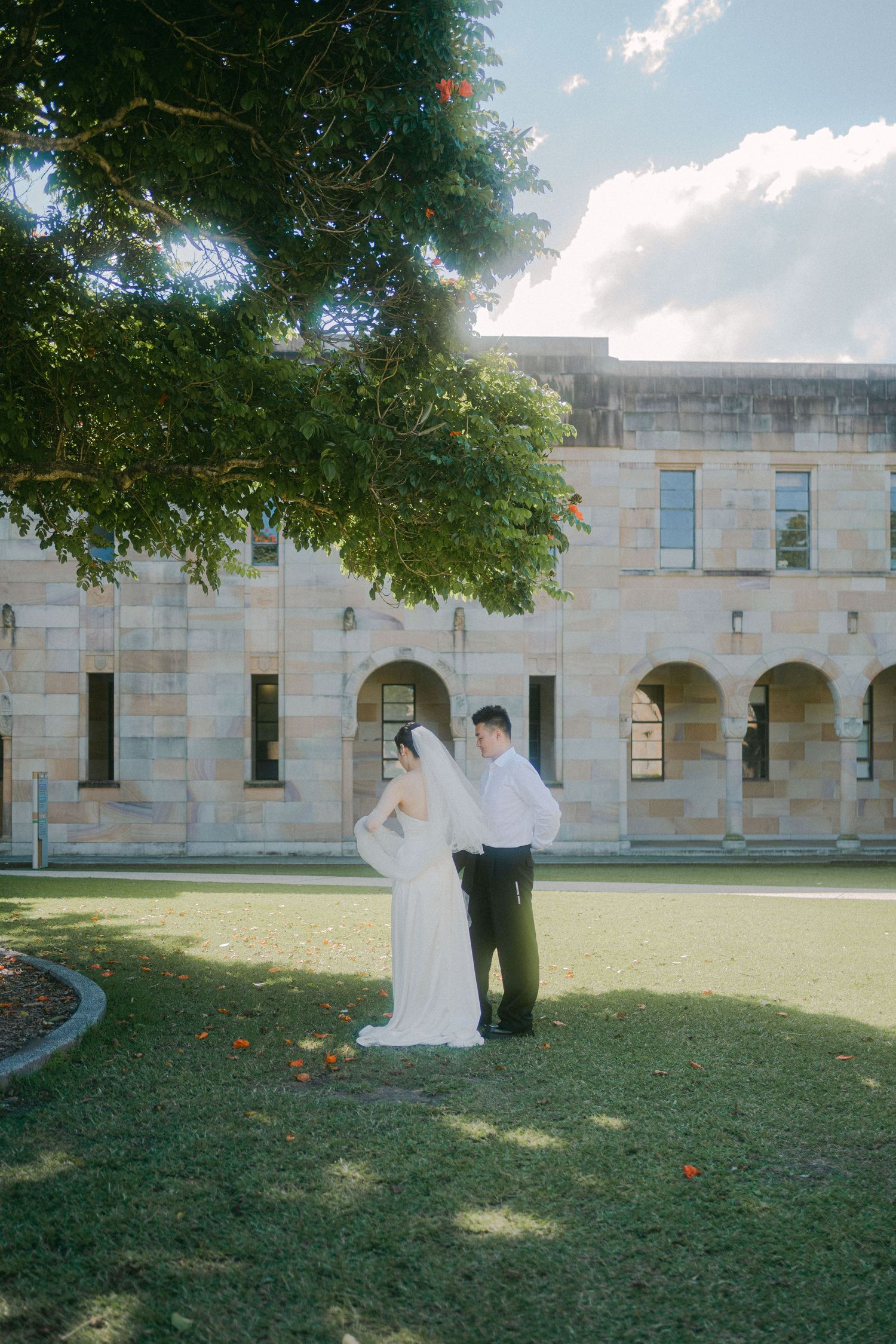 A wedding couple poses for photographs on the lawn in front of a grand stone building with arched architecture.