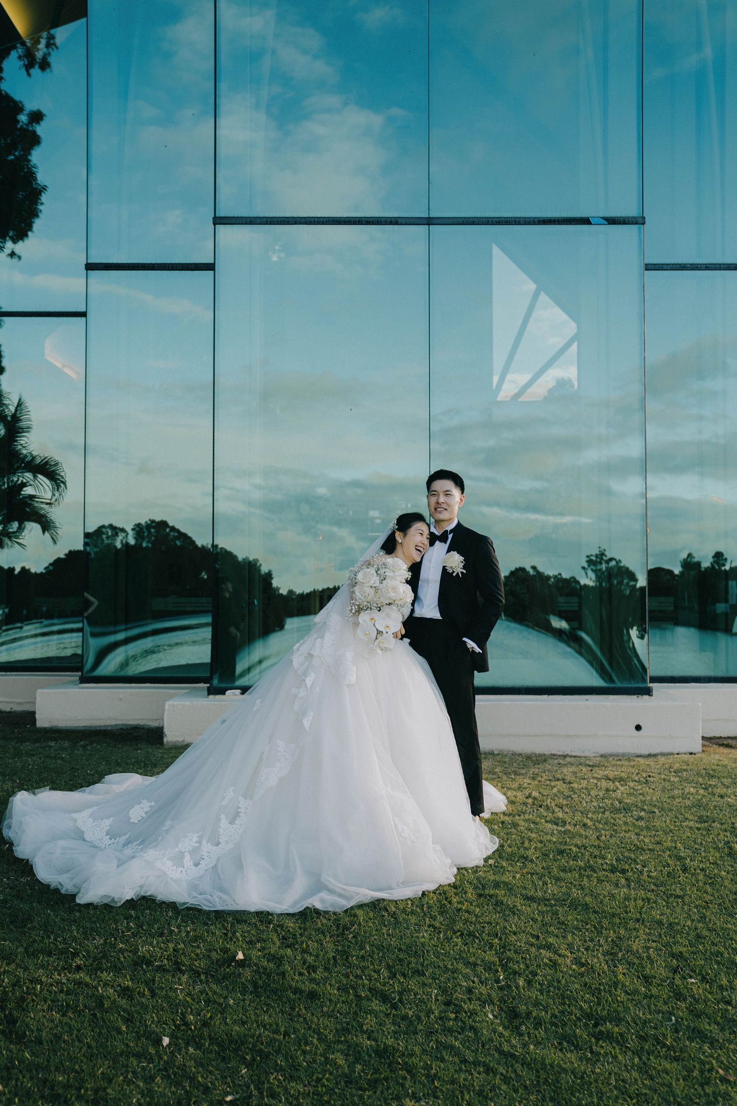 A couple in wedding attire poses in front of a modern glass building at sunset with a manicured lawn in the foreground.