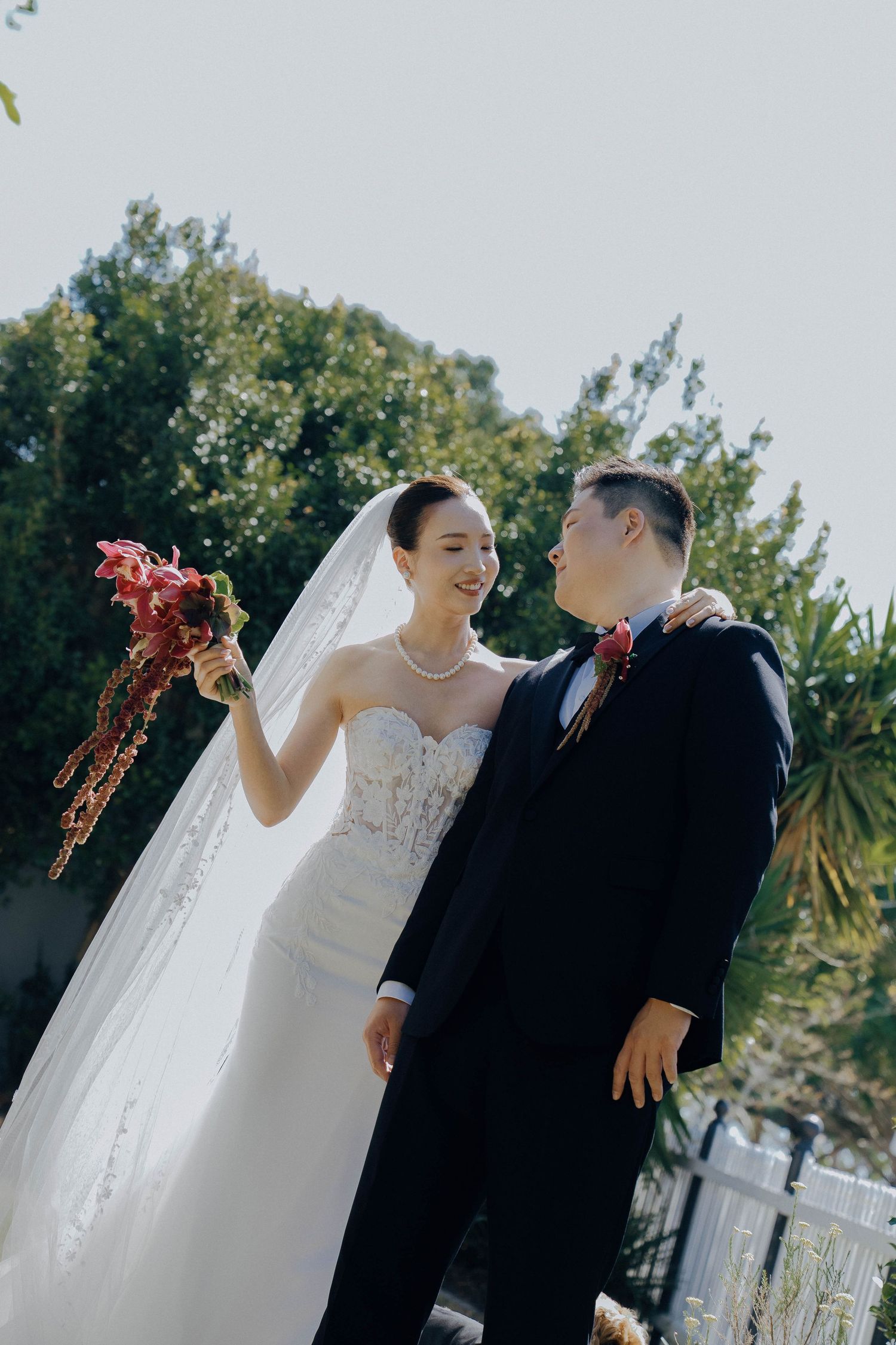 A wedding couple shares tender moments in a garden setting with palm trees and greenery in the background.