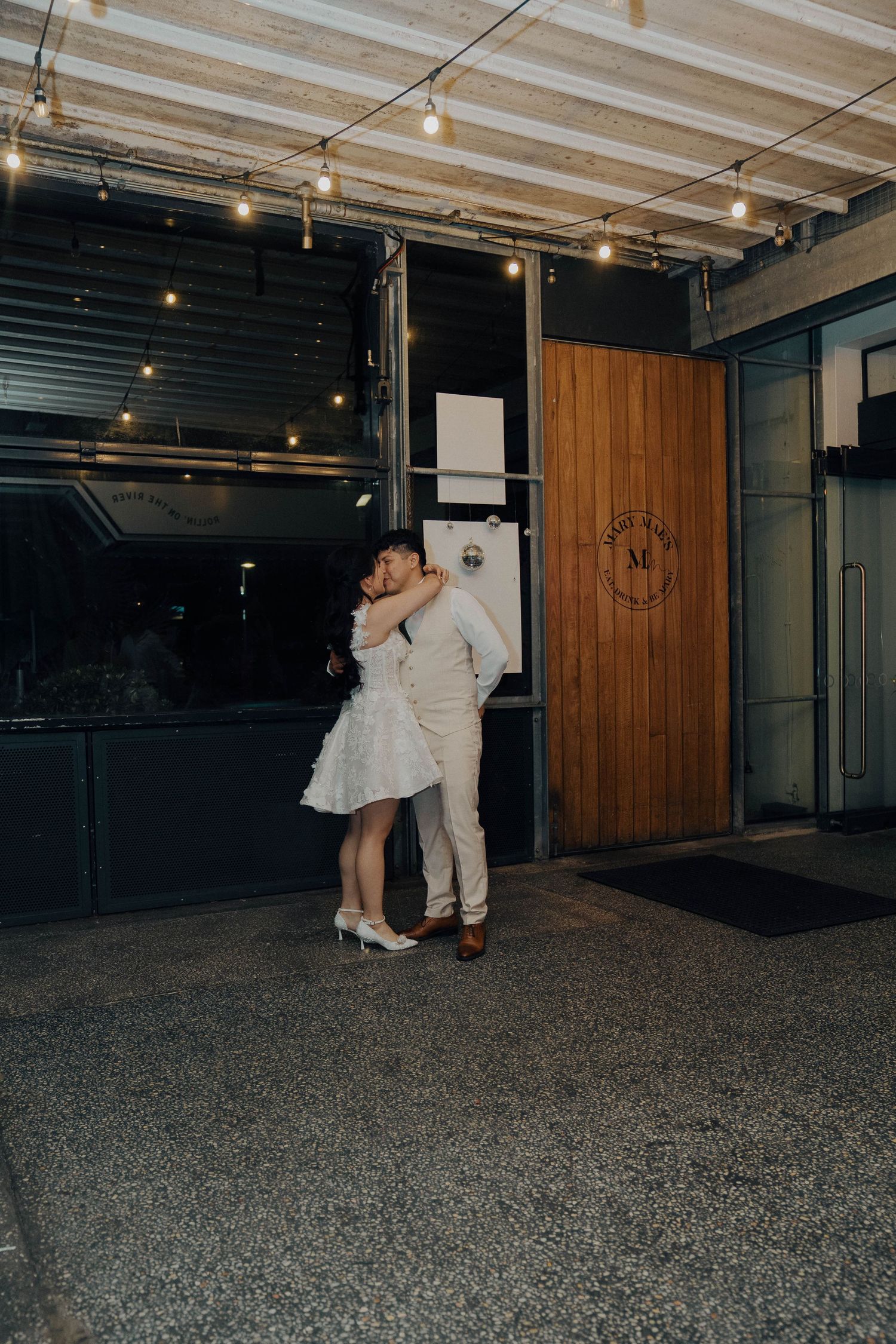 Two figures dance together in a dimly lit industrial space with exposed ceiling beams and string lights overhead.