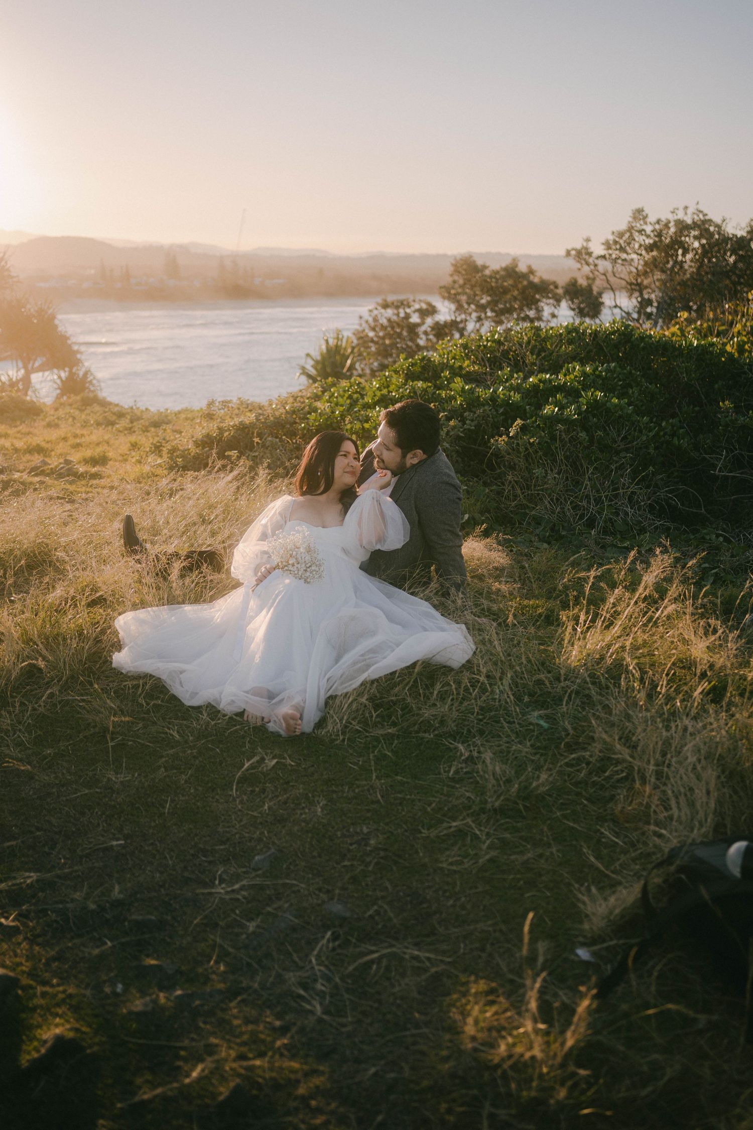 A couple in wedding attire sits together on a grassy coastal cliff at sunset, their white dress flowing in the breeze.