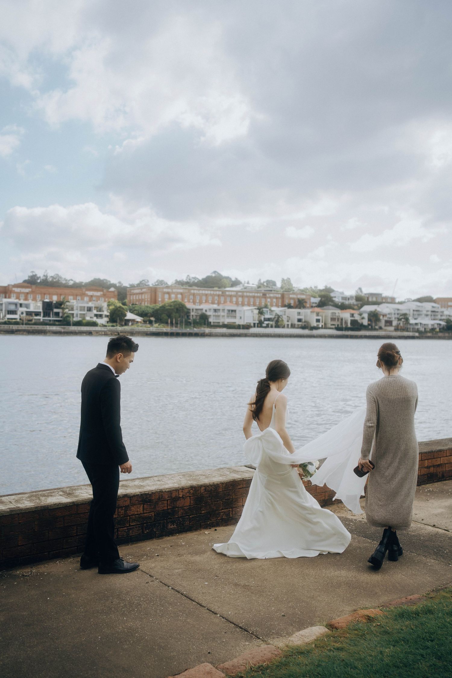 A wedding couple sits together on a waterfront promenade with buildings and cloudy skies in the background.