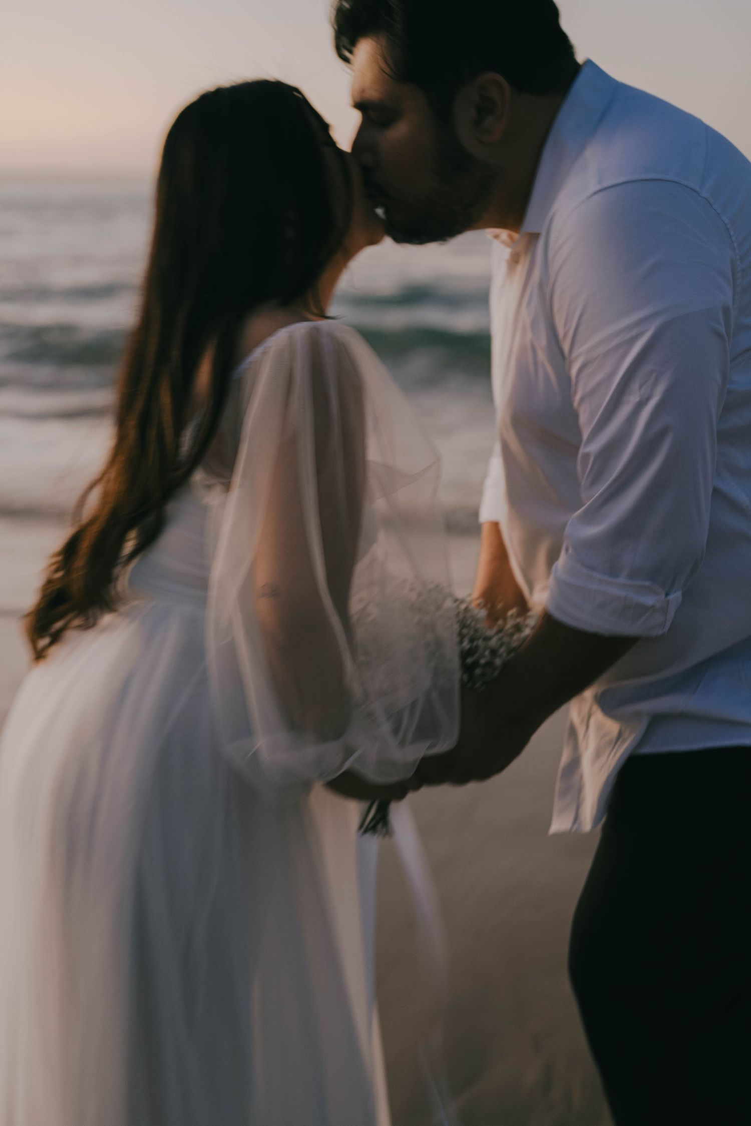 A romantic couple in white clothing embraces on a beach at sunset creating a dreamy silhouette effect.