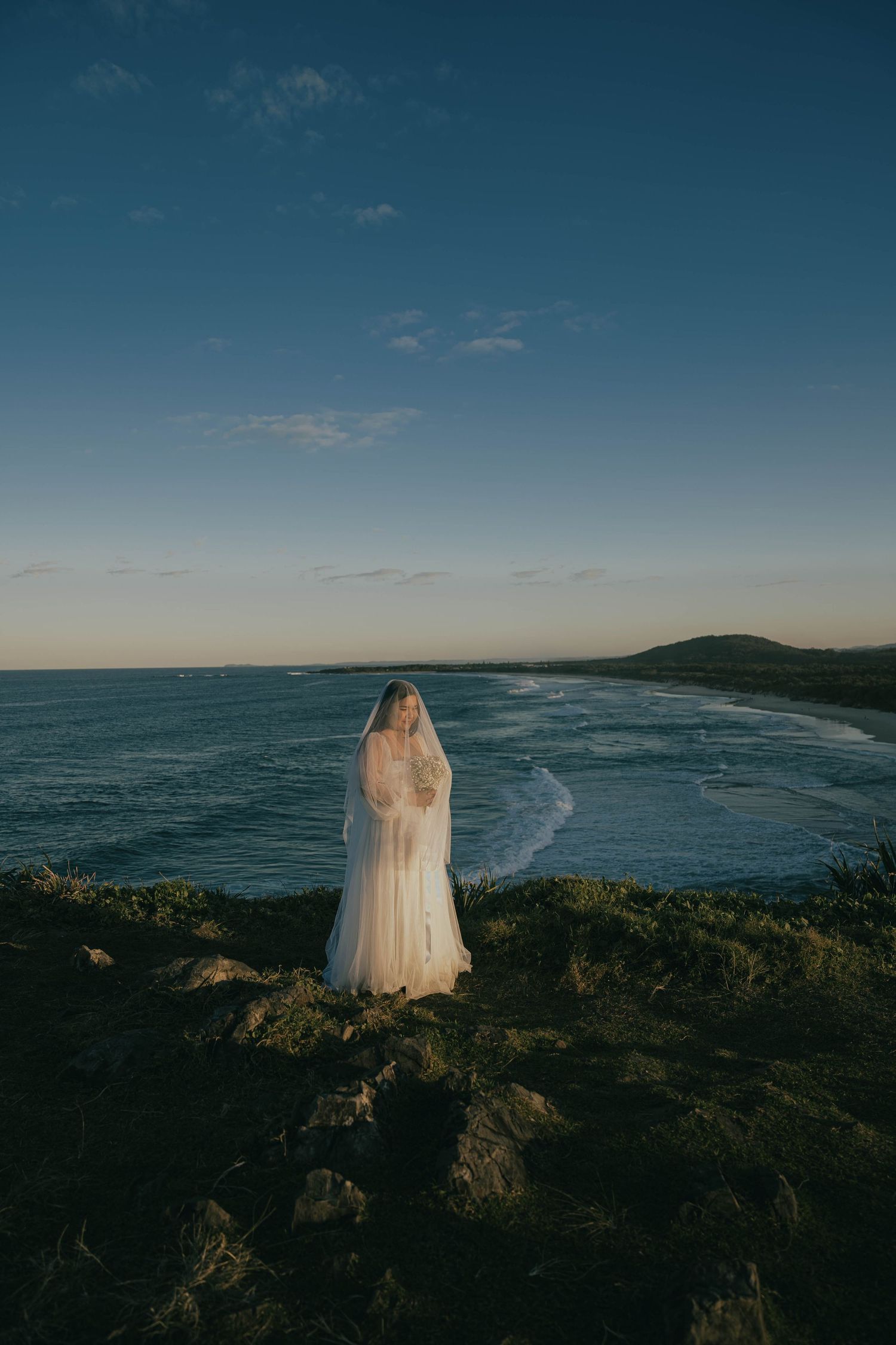 A panoramic coastal view at sunset shows a white-clad figure standing on rocky cliffs overlooking a dramatic ocean shoreline.