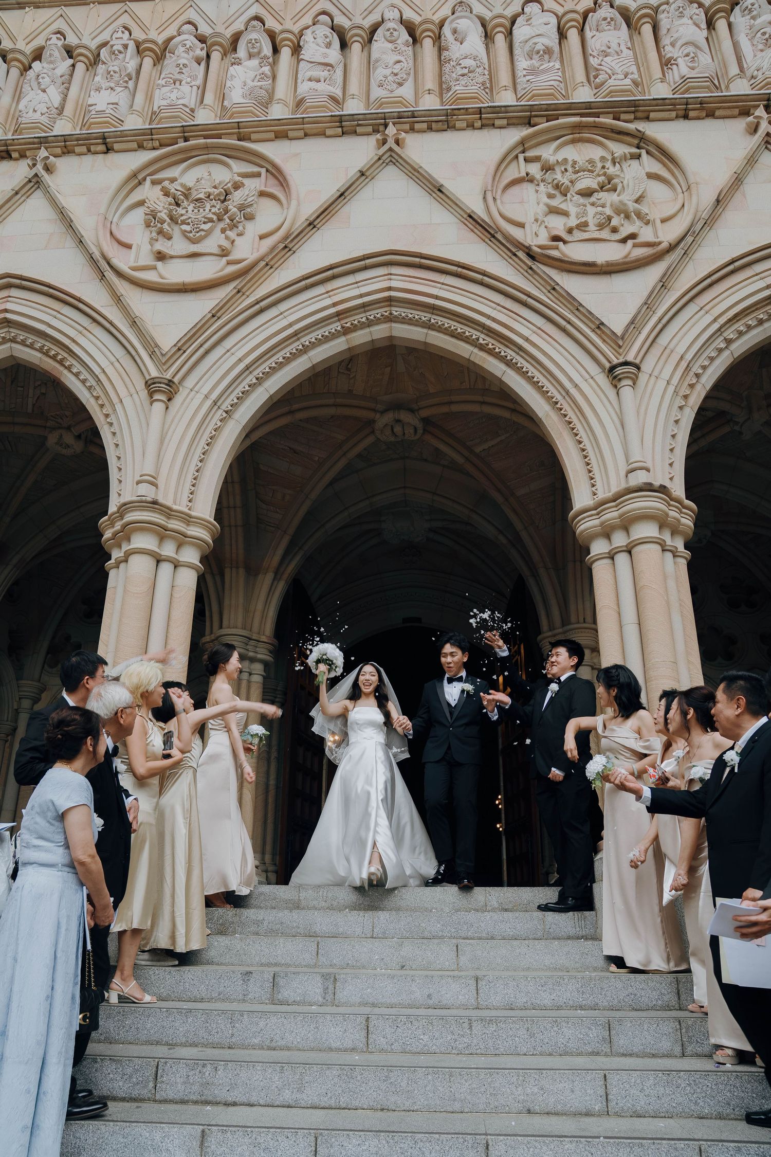Wedding party gathers on ornate stone church steps with Gothic architectural details and arched doorways.
