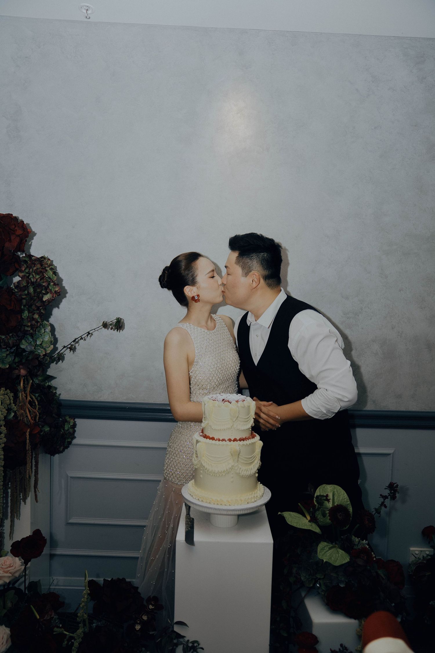 A couple shares a romantic kiss behind their elegant two-tier white wedding cake at their reception.