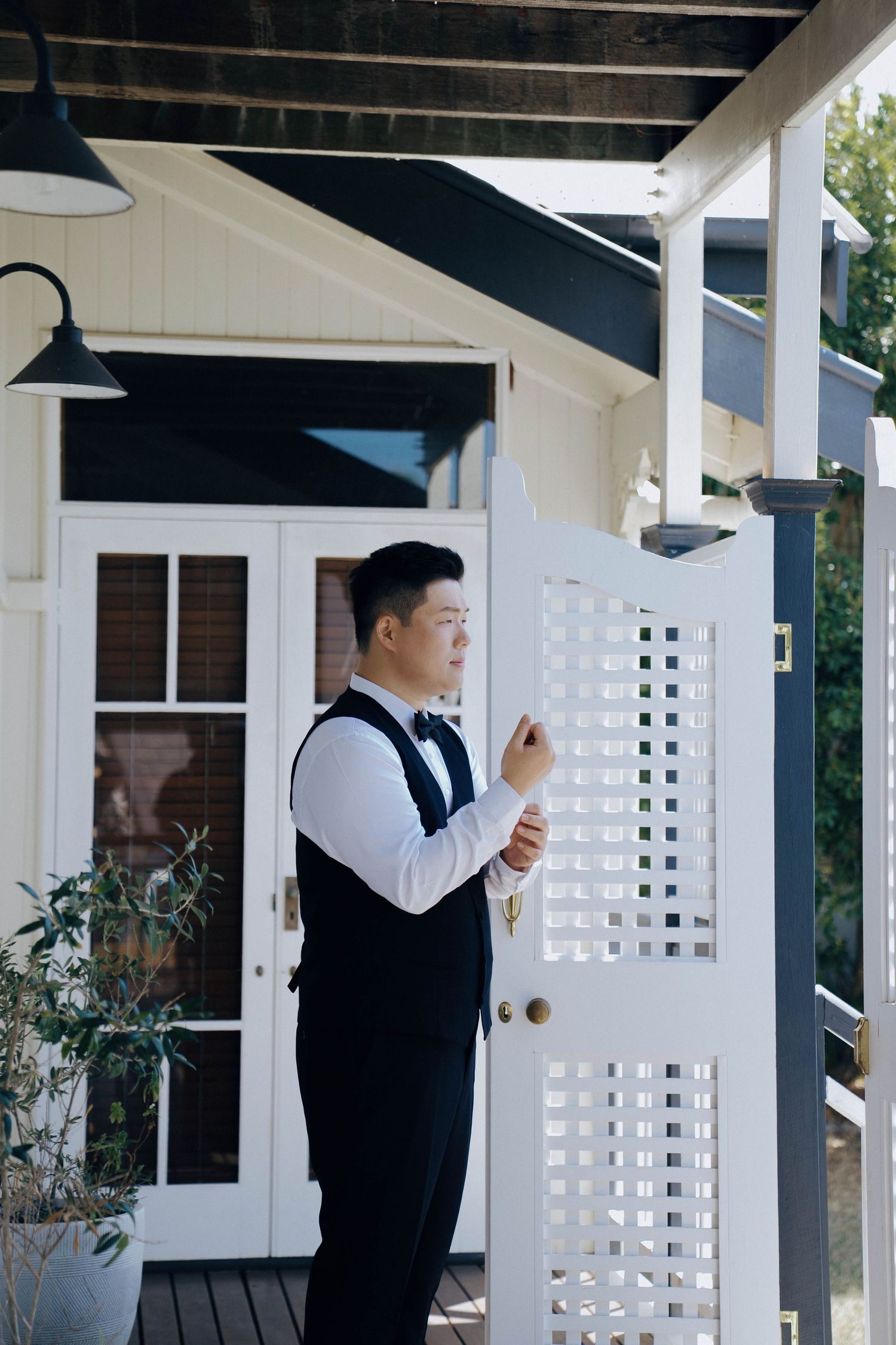 Two people in formal attire stand on an elegant white porch with decorative lattice panels and columns.
