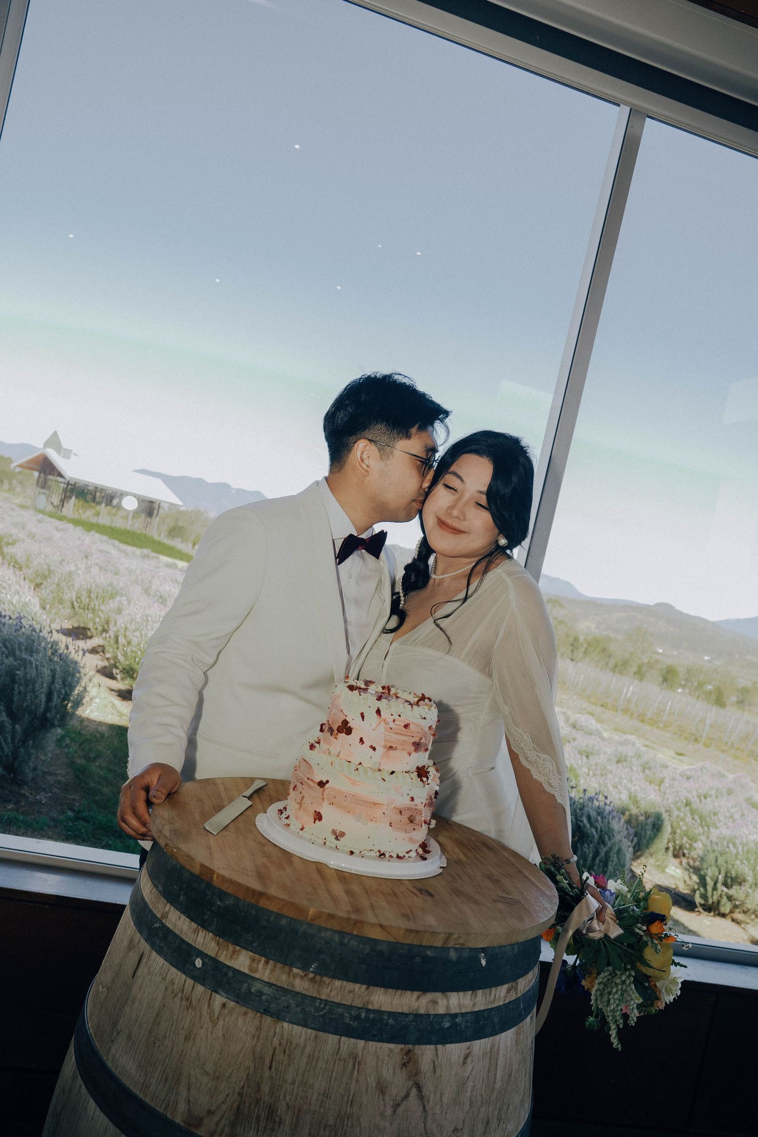 A romantic couple shares an intimate moment while posing with a rustic wine barrel against a scenic mountain backdrop.