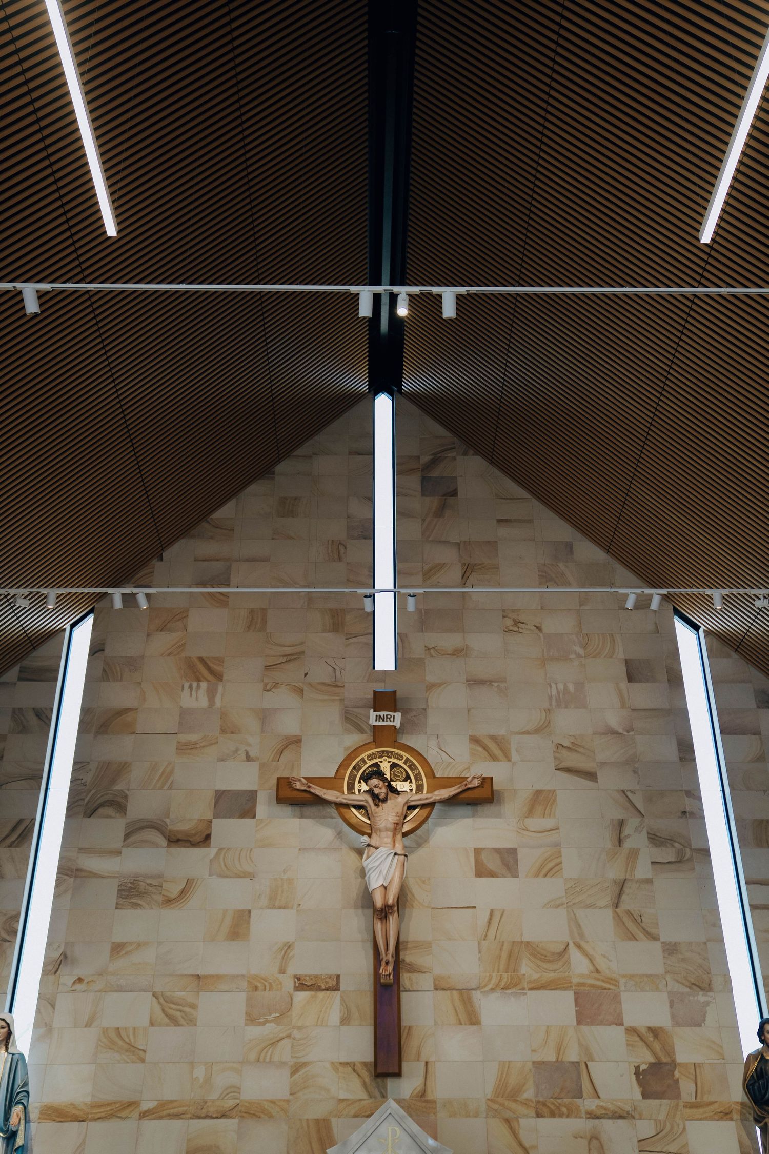 A crucifix mounted on a beige stone wall under modern lighting in a church with angular wooden ceiling panels.