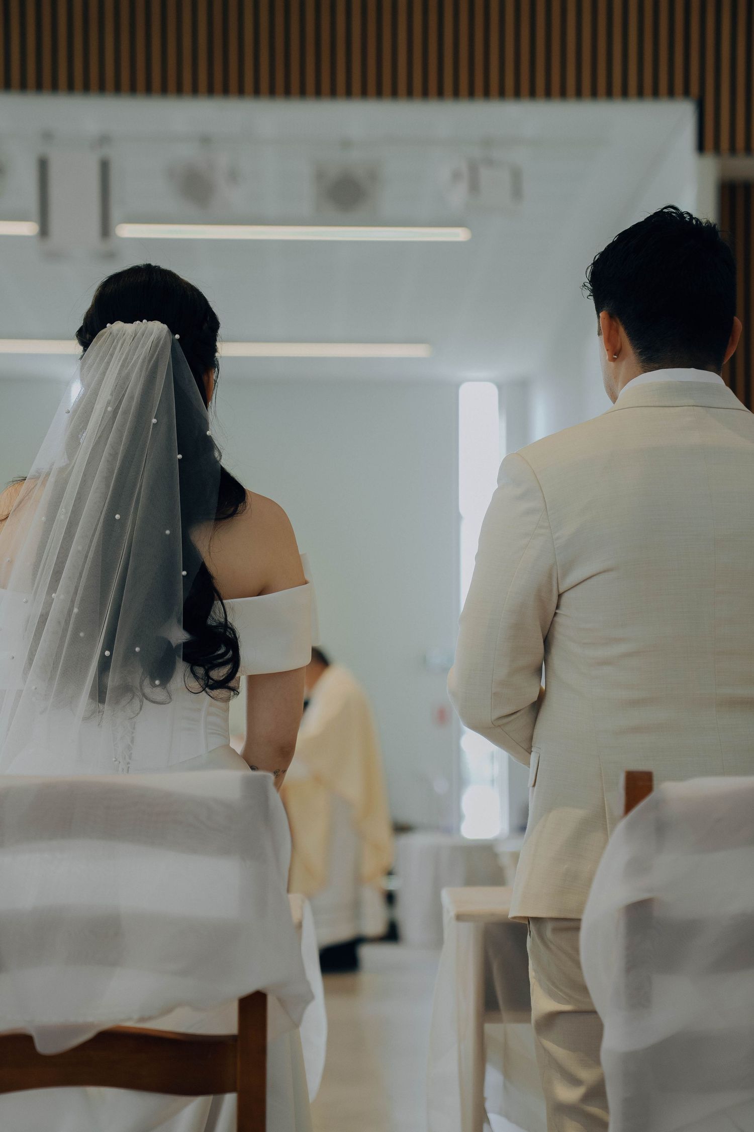 A couple in white wedding attire sits together at a table during their wedding ceremony.
