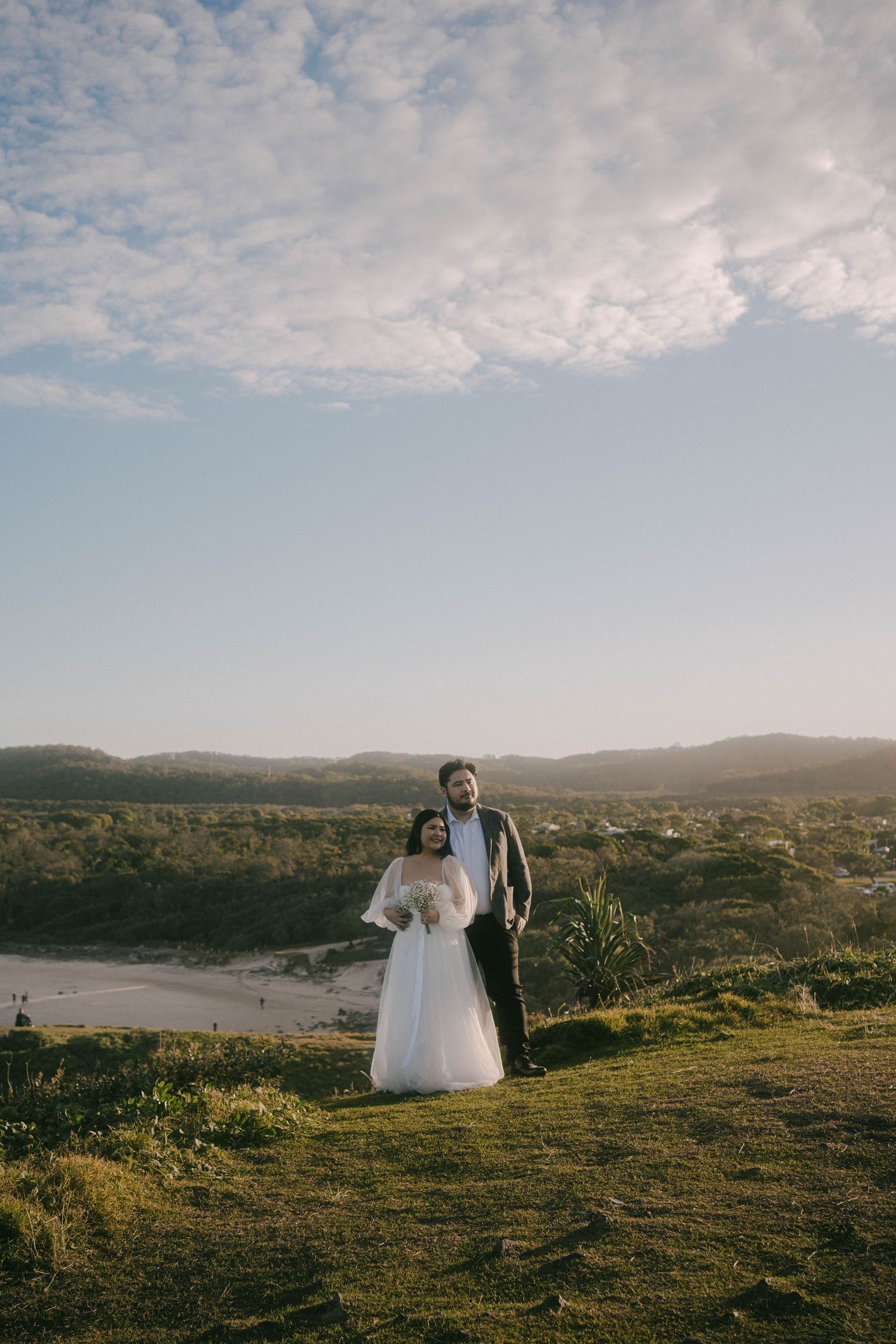 A couple in wedding attire stands on a grassy hilltop overlooking a scenic coastal landscape at sunset.