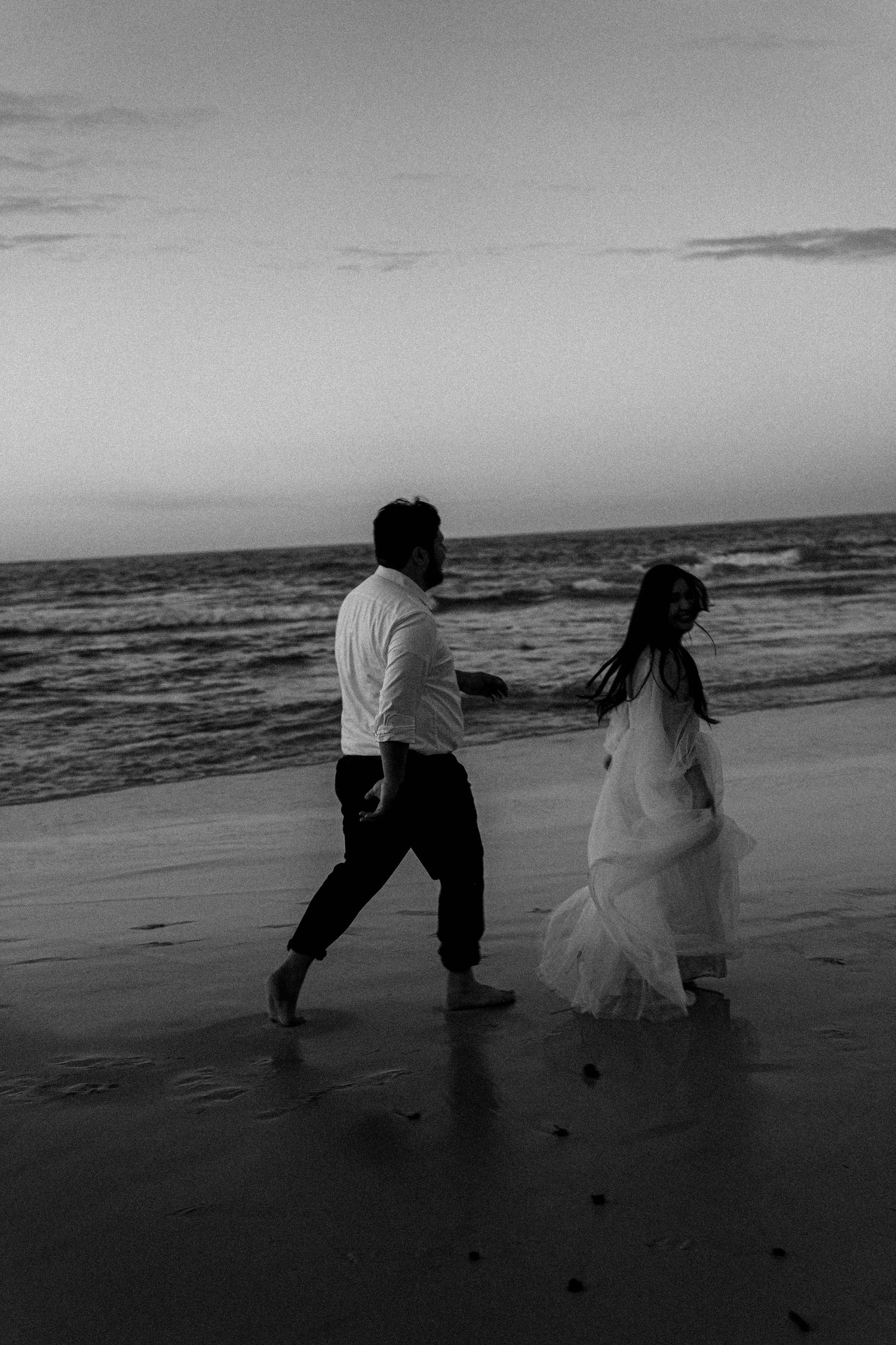 A couple in wedding attire walks along a misty beach at sunset, captured in black and white photography.