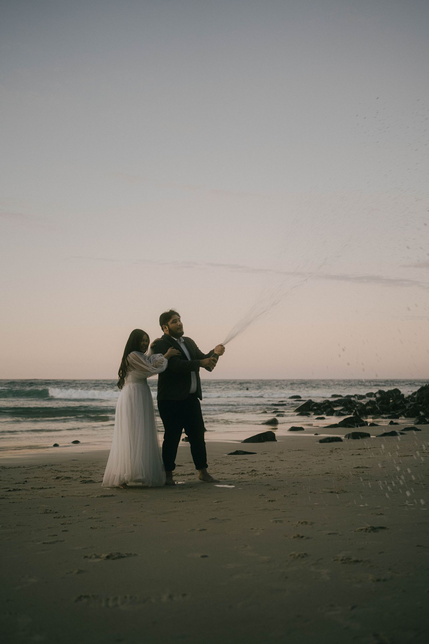 A couple in wedding attire walks along a misty beach at dusk, creating a romantic silhouette against the ocean waves.