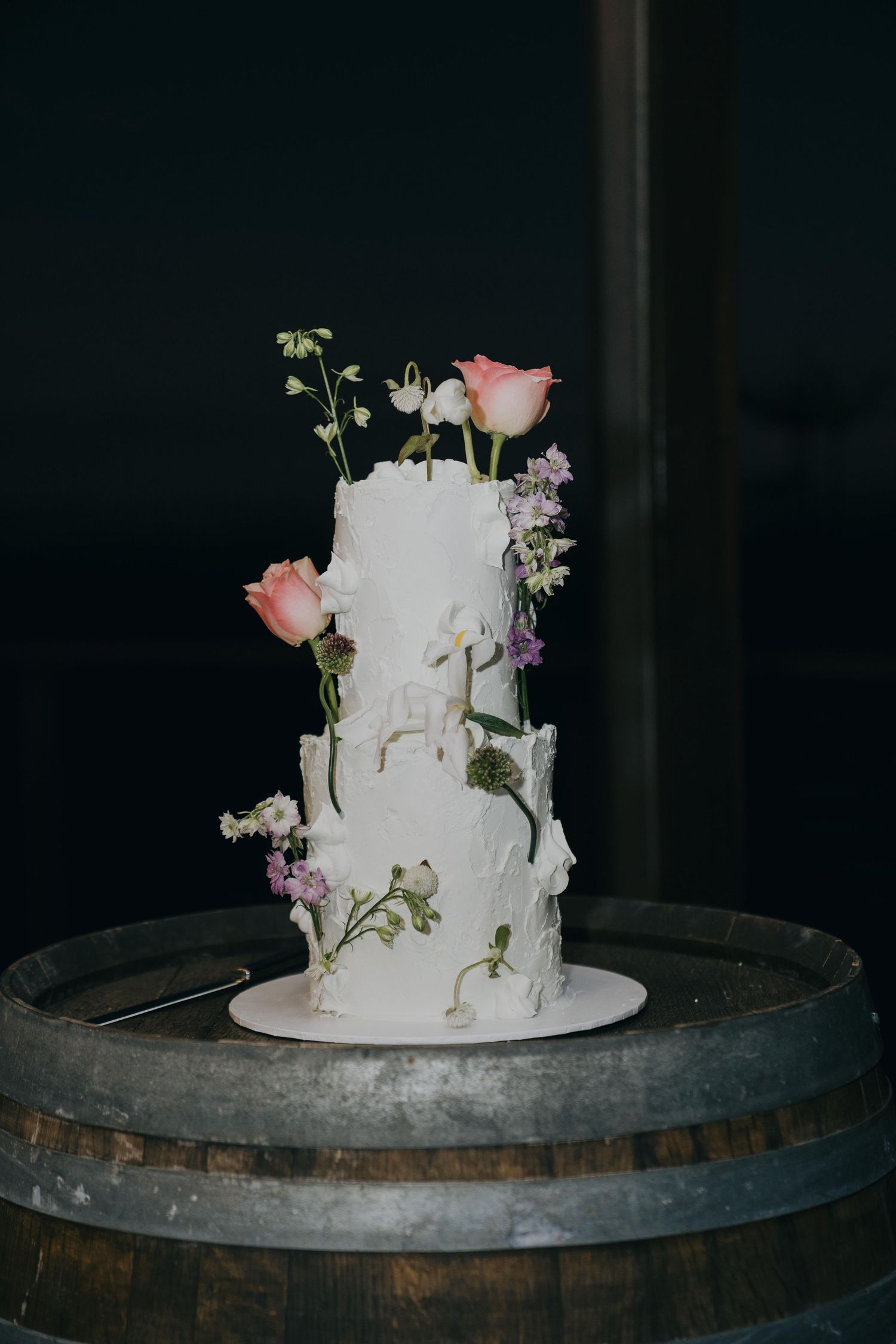 A rustic white wedding cake decorated with fresh flowers sits atop a wooden barrel in a dark moody setting.