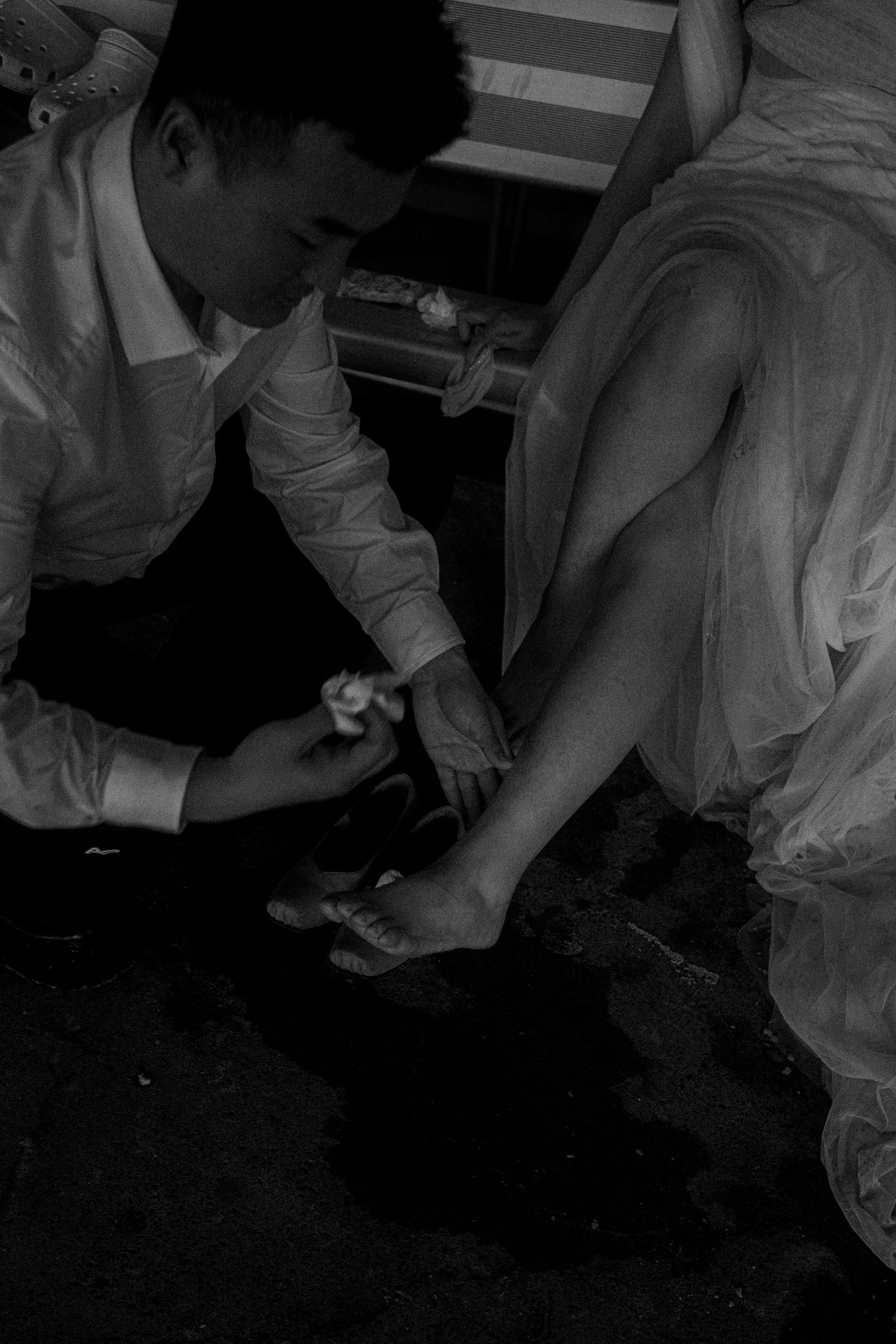 A black and white photo shows a groom placing a decorative shoe on a bride's foot in an intimate wedding moment.