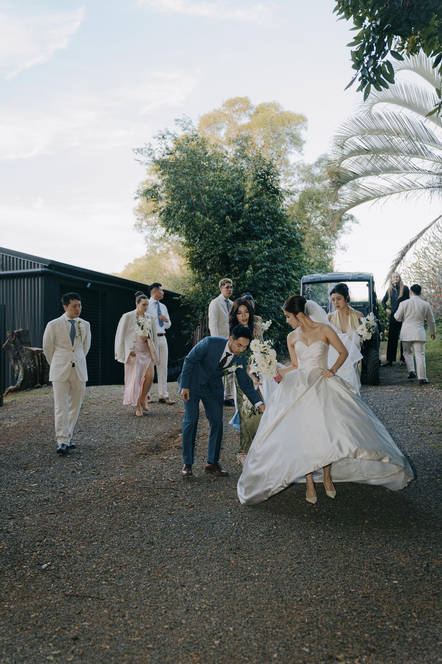 A wedding party in formal attire helps the bride navigate across a hilly outdoor terrain during a photo session.
