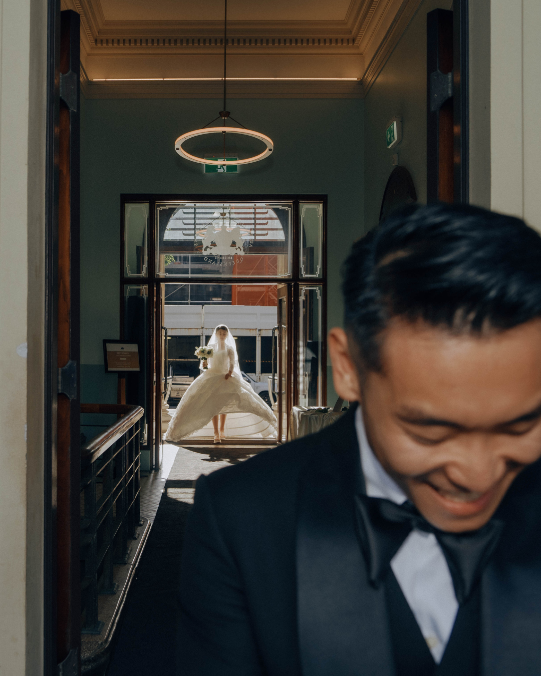 A person in a tuxedo smiling near a doorway while a blurred white wedding dress figure appears in the background.